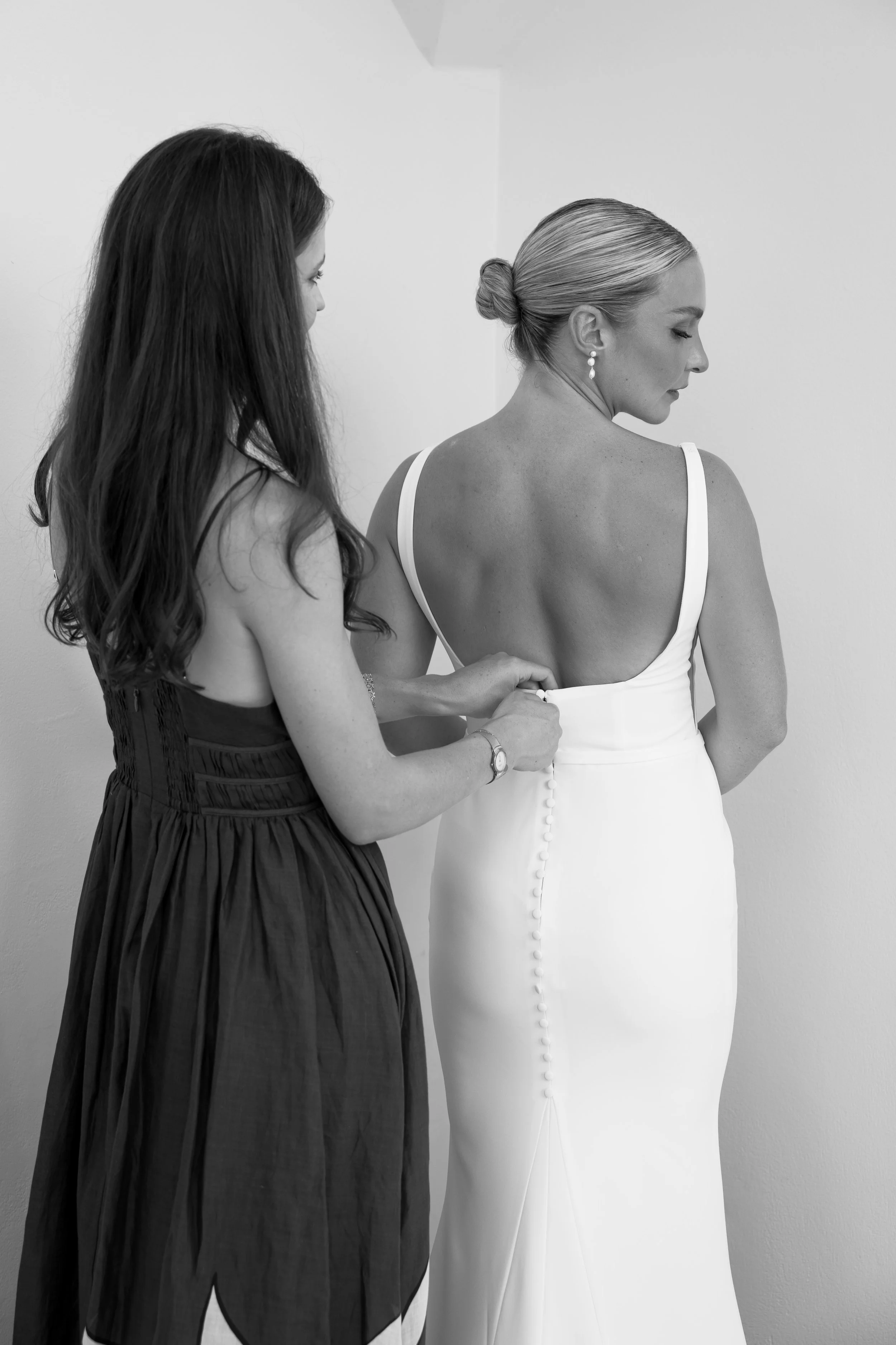 A woman in a white dress getting help buttoning it from another woman in a black dress