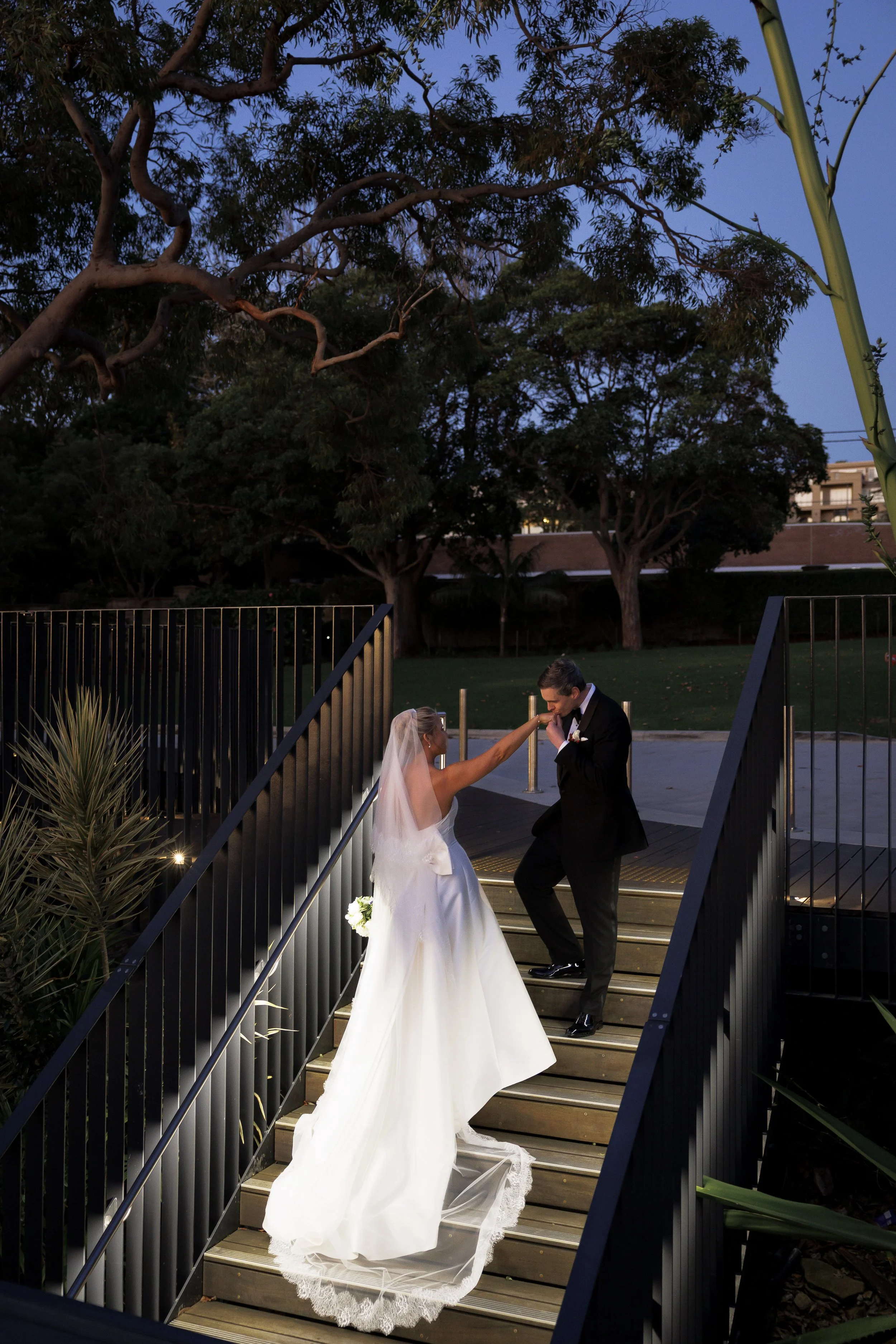 A bride and groom sharing a dance on an outdoor staircase during a wedding, with trees and a park in the background at dusk.