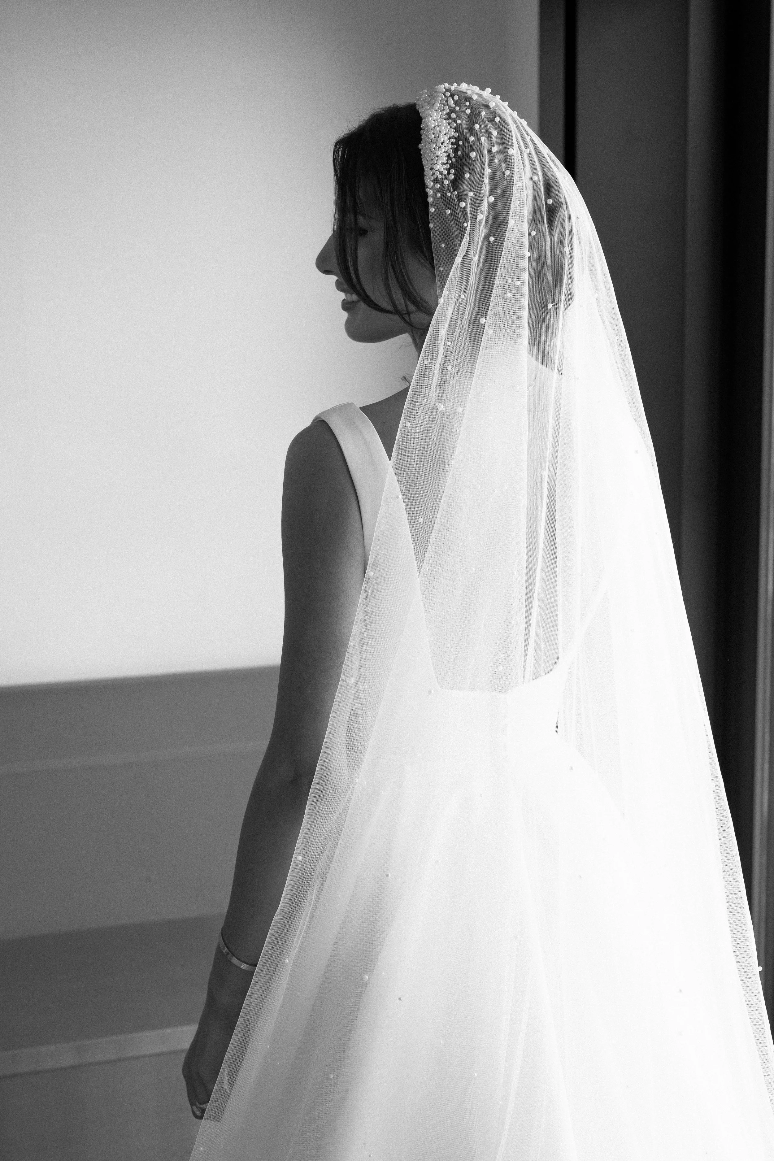 A woman wearing a wedding dress and veil, smiling and looking to the side indoors.