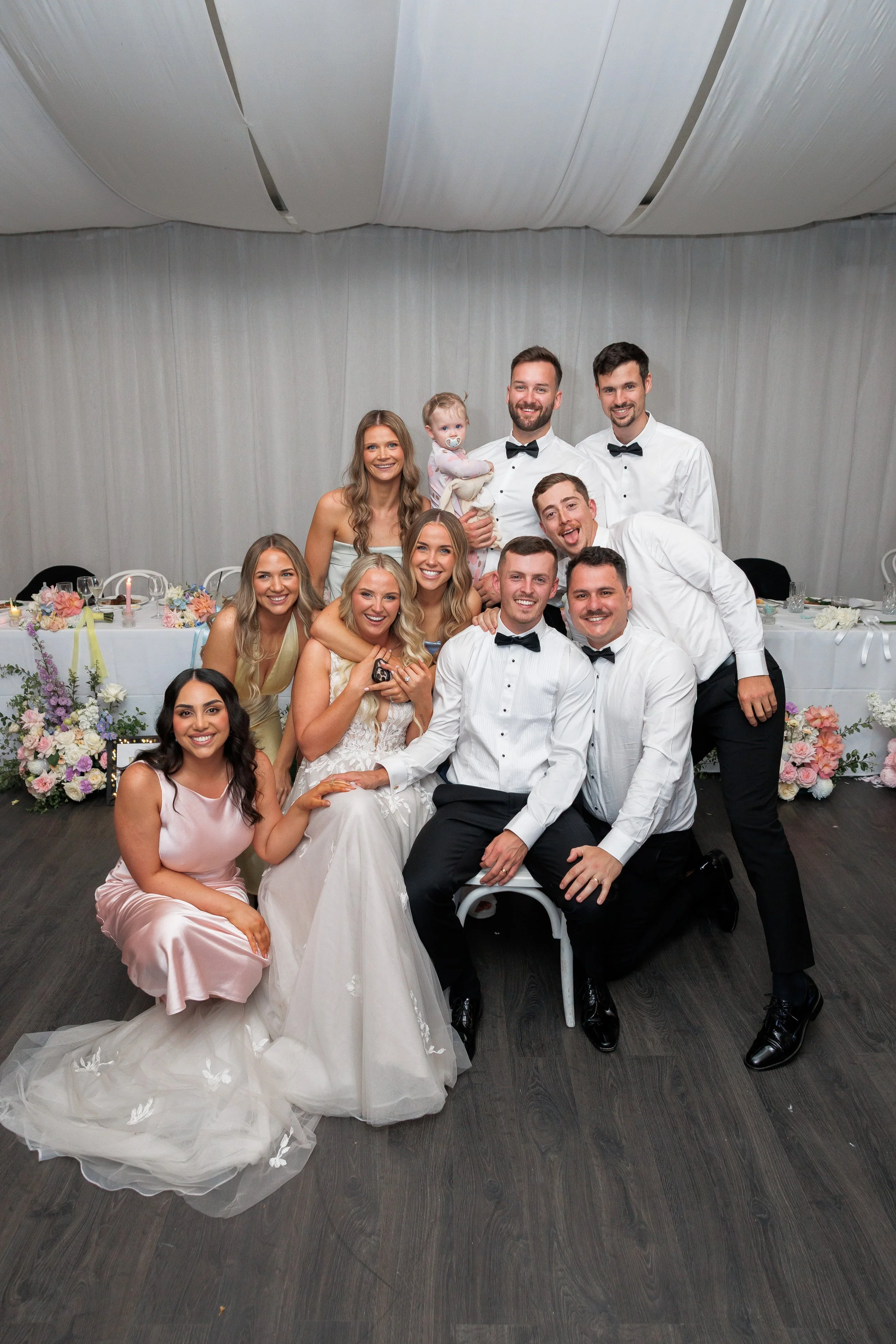 A group of people at a wedding reception, including the bride and groom, family, friends, and a young child, all smiling together in a decorated event space.
