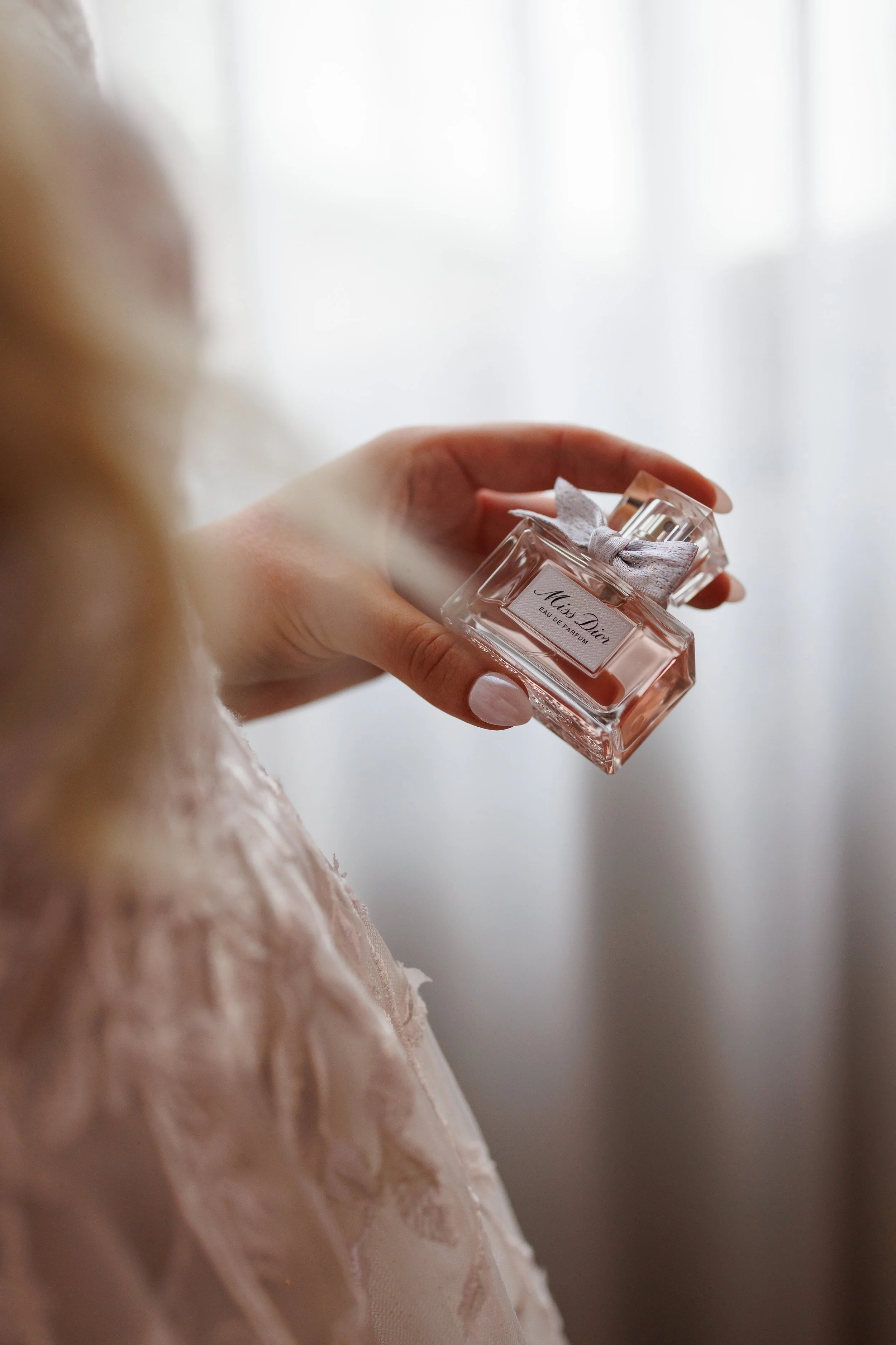 A woman holding a bottle of Miss Dior perfume with a winged ribbon bow on the cap, in front of a softly lit curtain.