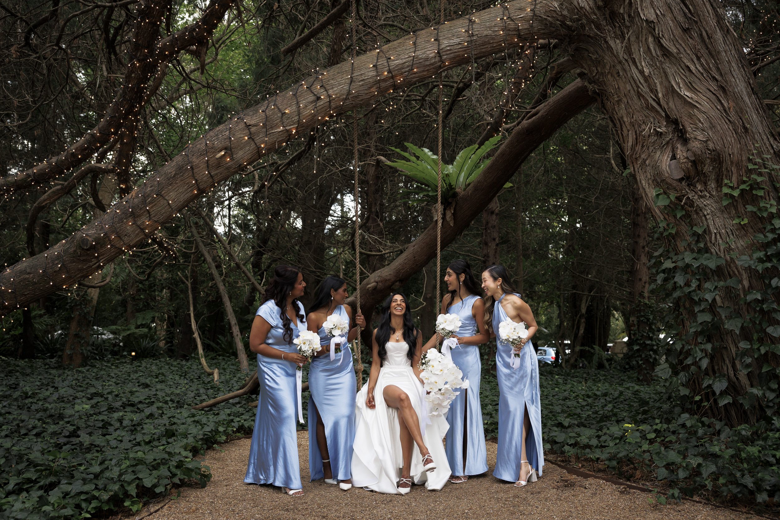 A bride in a white wedding dress seated on a swing, surrounded by five bridesmaids in matching light blue dresses, holding white bouquets, in a forest setting with string lights on the trees.