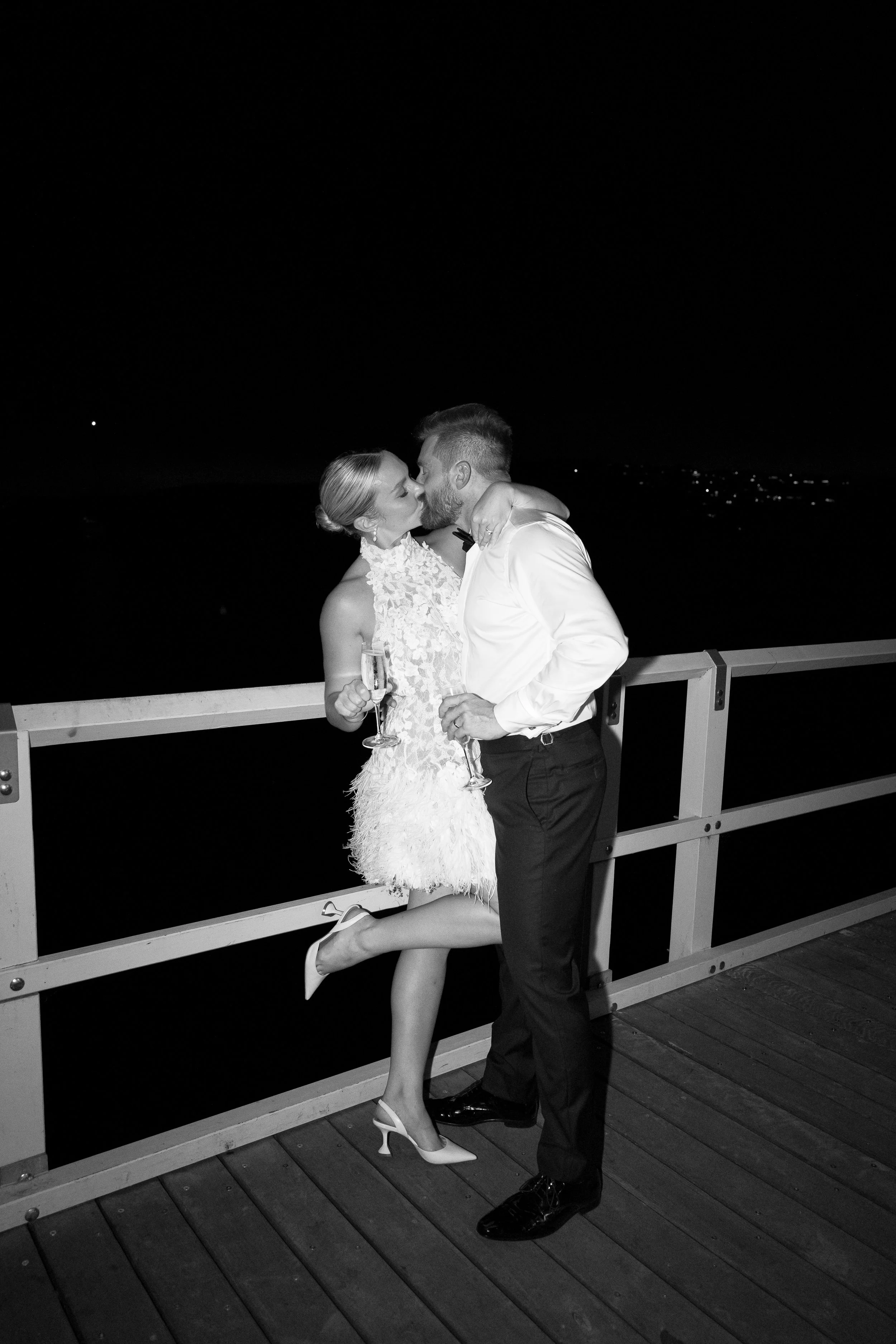 A couple kissing on a pier at night, holding champagne glasses, with a dark sky and distant city lights in the background.