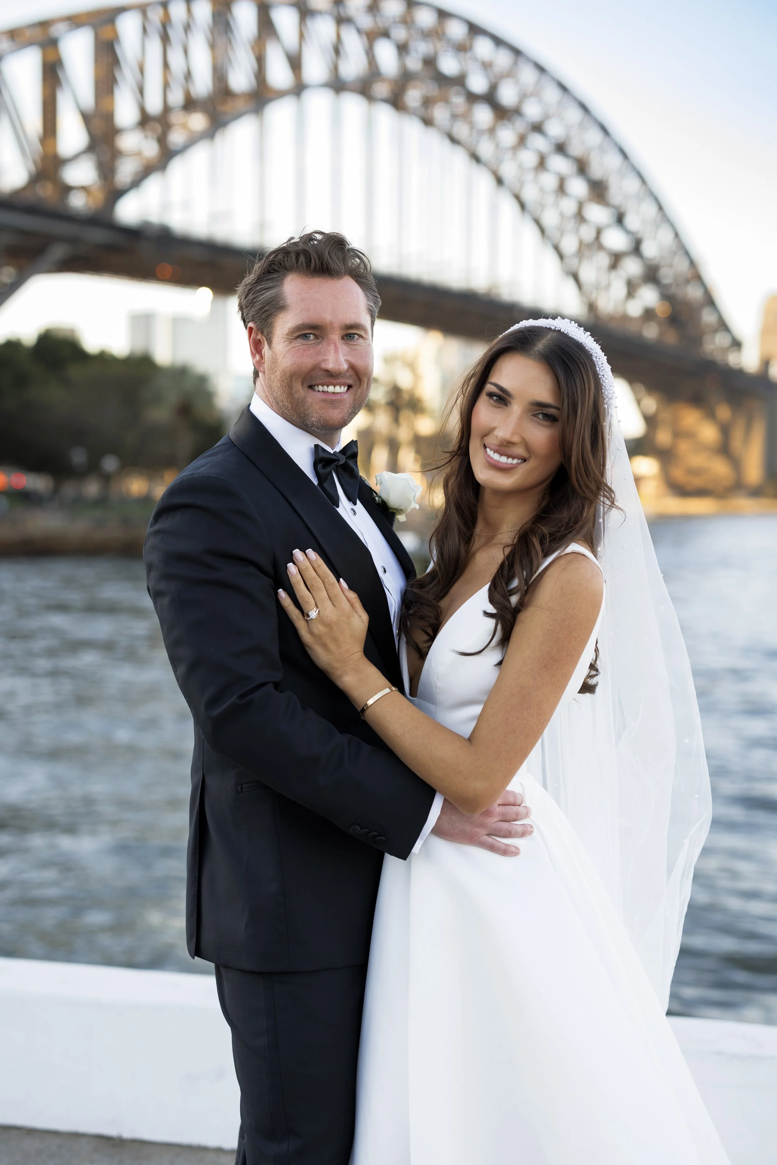 A newlywed couple smiling and posing together outdoors near a body of water, with a large bridge in the background at sunset.