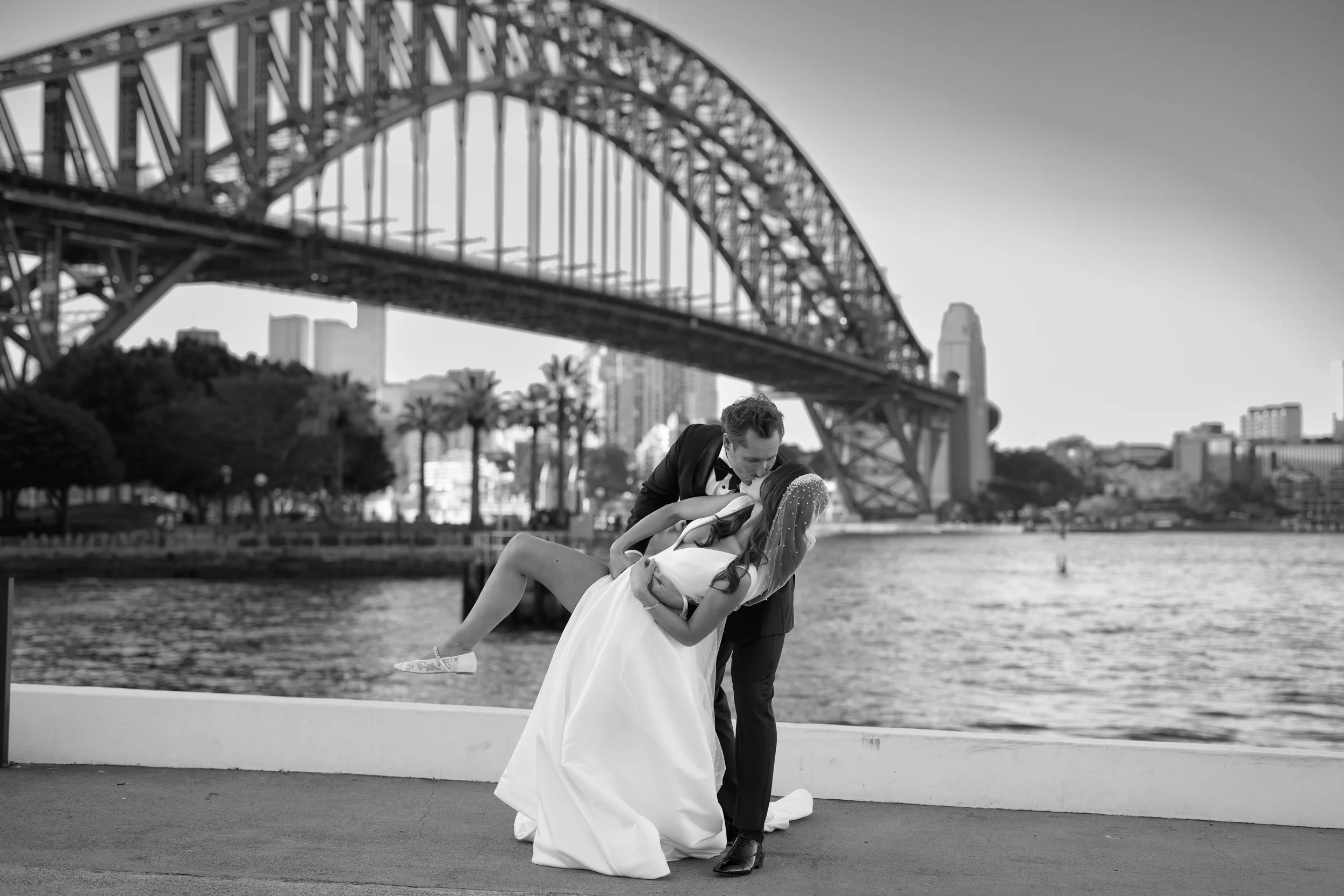A black and white photo of a couple (bride and groom) by a waterfront, with a large bridge and city skyline in the background. The groom is dipping the bride during a kiss.