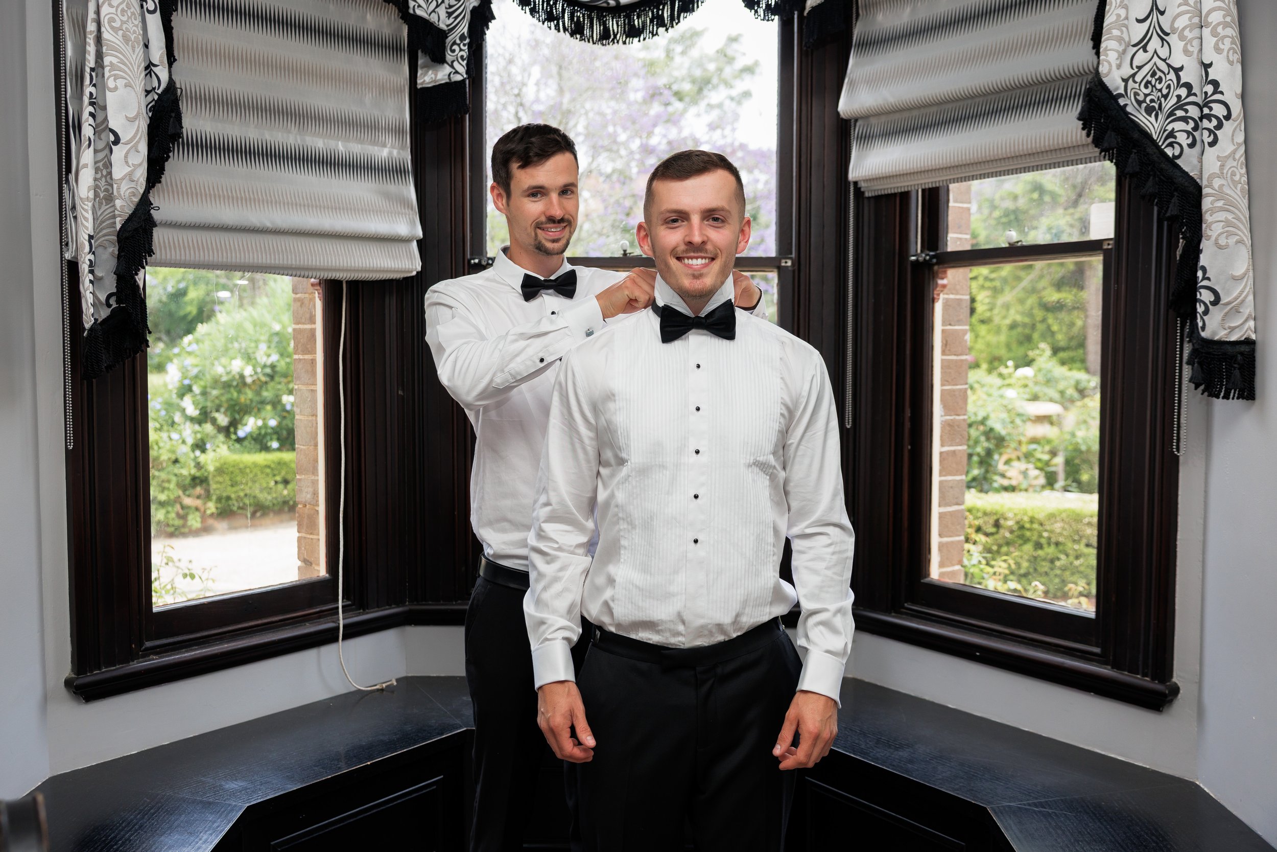 Two men in formal attire, one helping the other adjust his black bow tie, standing in front of a bay window with patterned curtains and lush garden outside.
