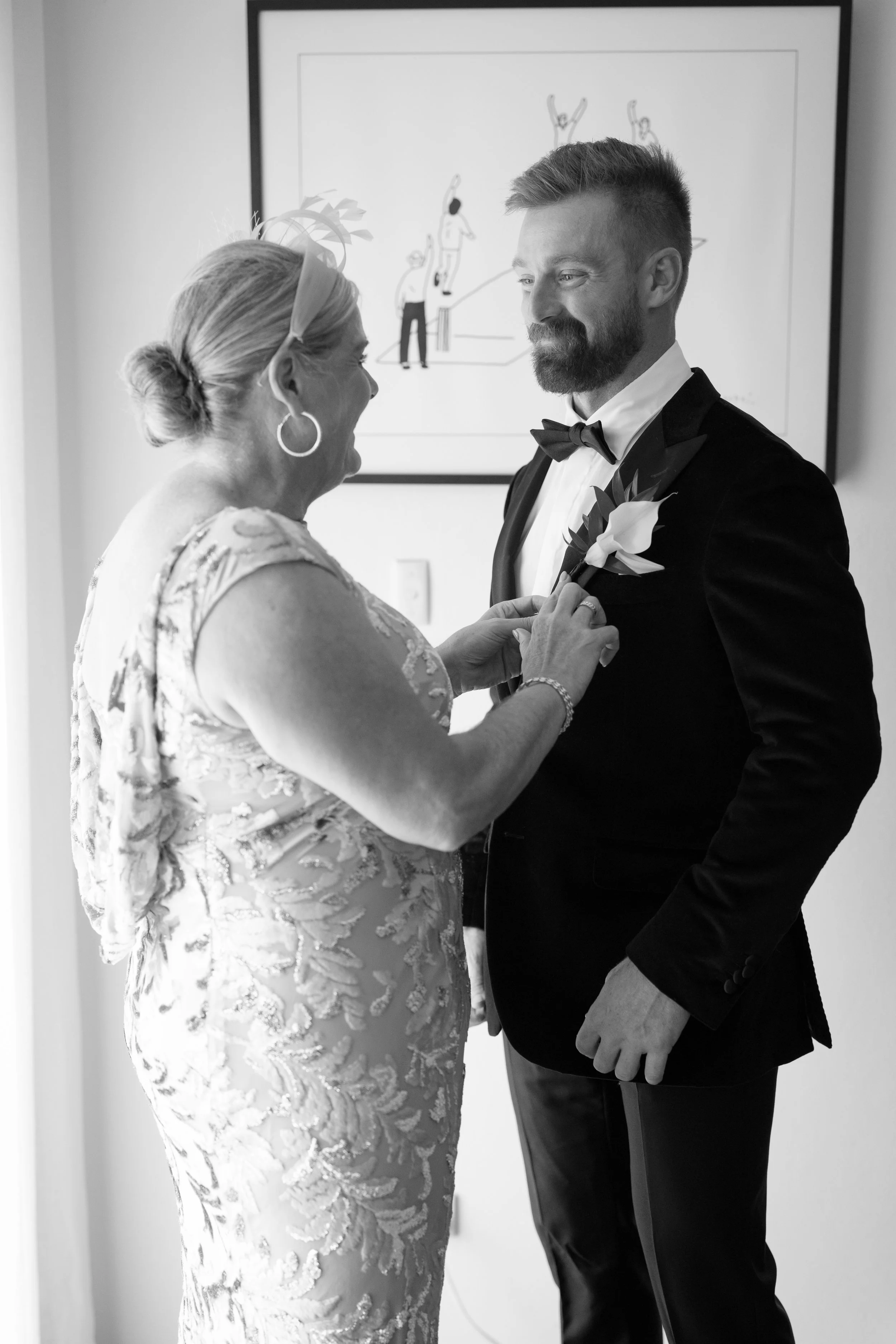 A woman dressed in a lace gown pins a flower onto a man's tuxedo in a wedding setting, with artwork on the wall behind them.