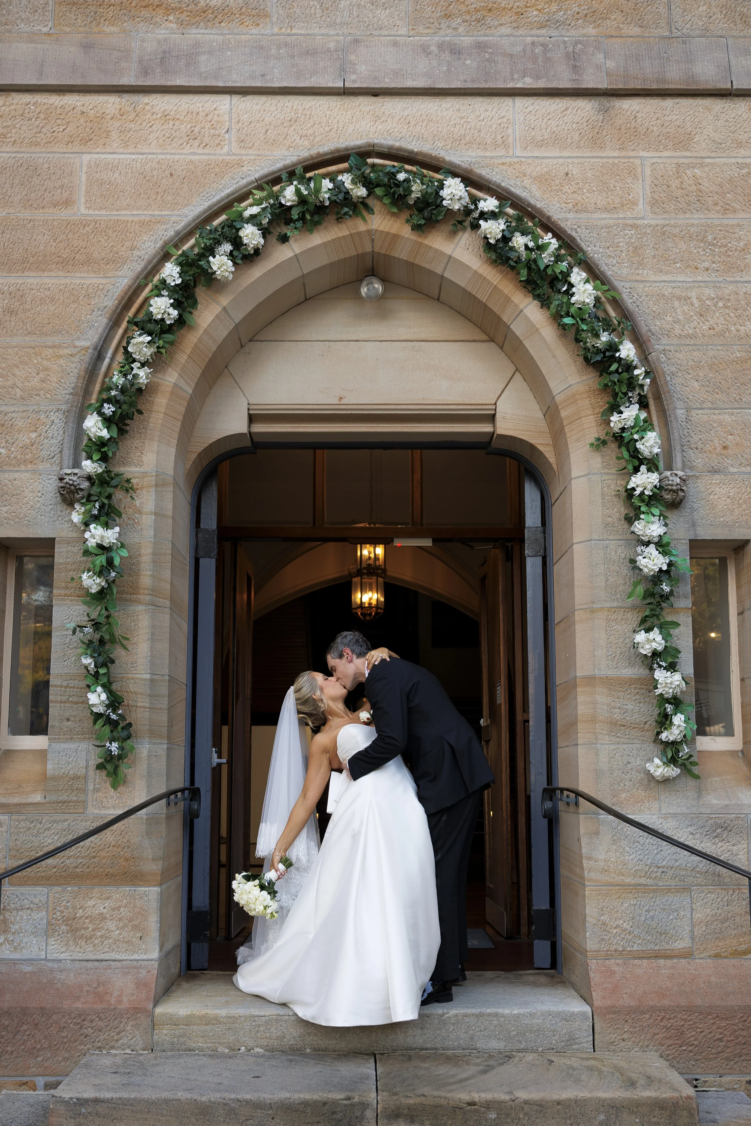 A bride and groom share a kiss on the steps of a church decorated with a floral archway.