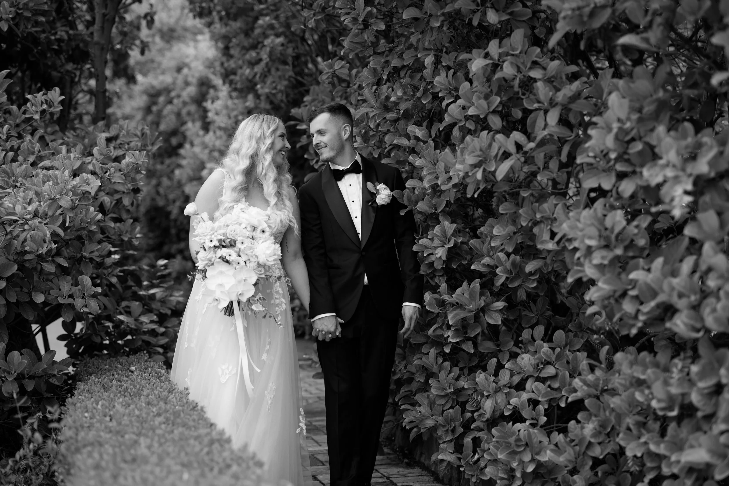 A bride and groom walk hand in hand through a garden, smiling at each other. The bride has long, wavy hair and wears a white wedding dress with floral details, holding a bouquet. The groom is dressed in a black tuxedo with a bow tie and a boutonniere. They are surrounded by lush foliage.