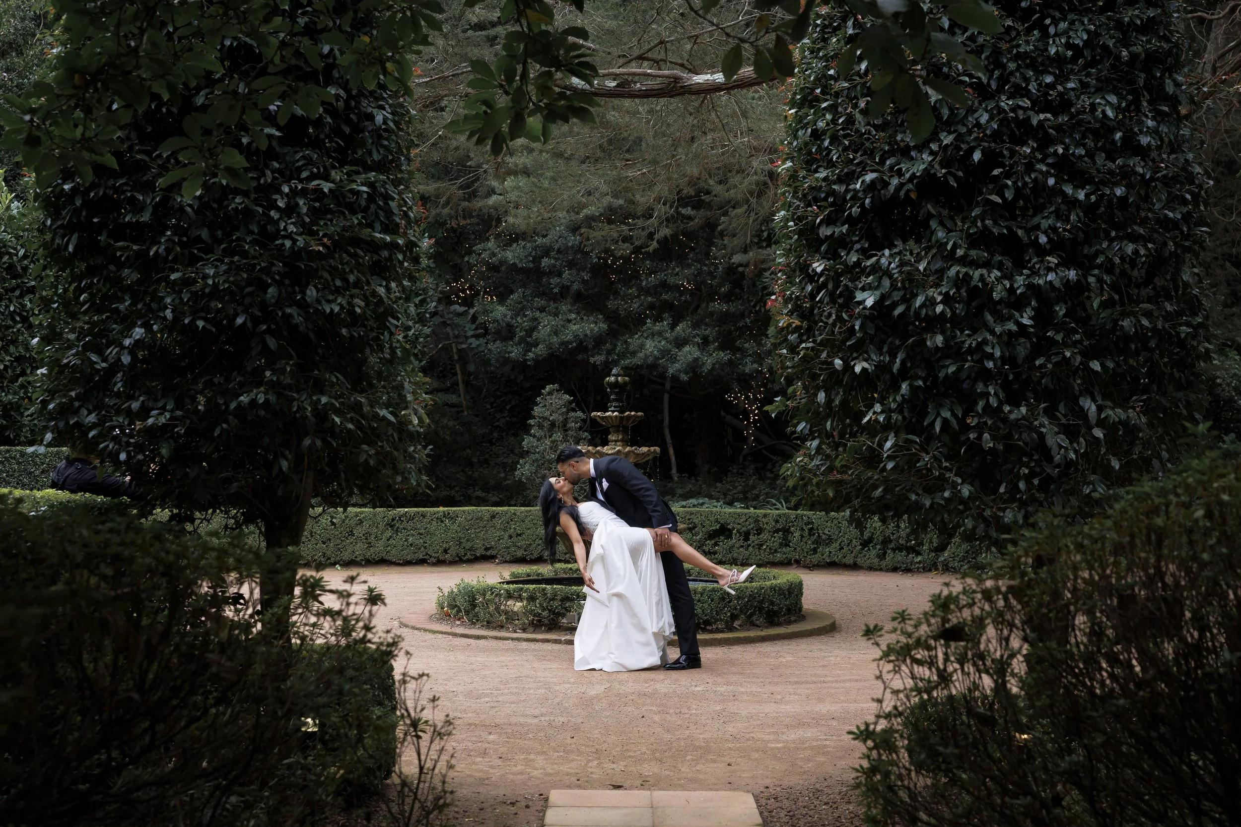 A newlywed couple sharing a kiss in a garden with a fountain, surrounded by lush hedge and trees.