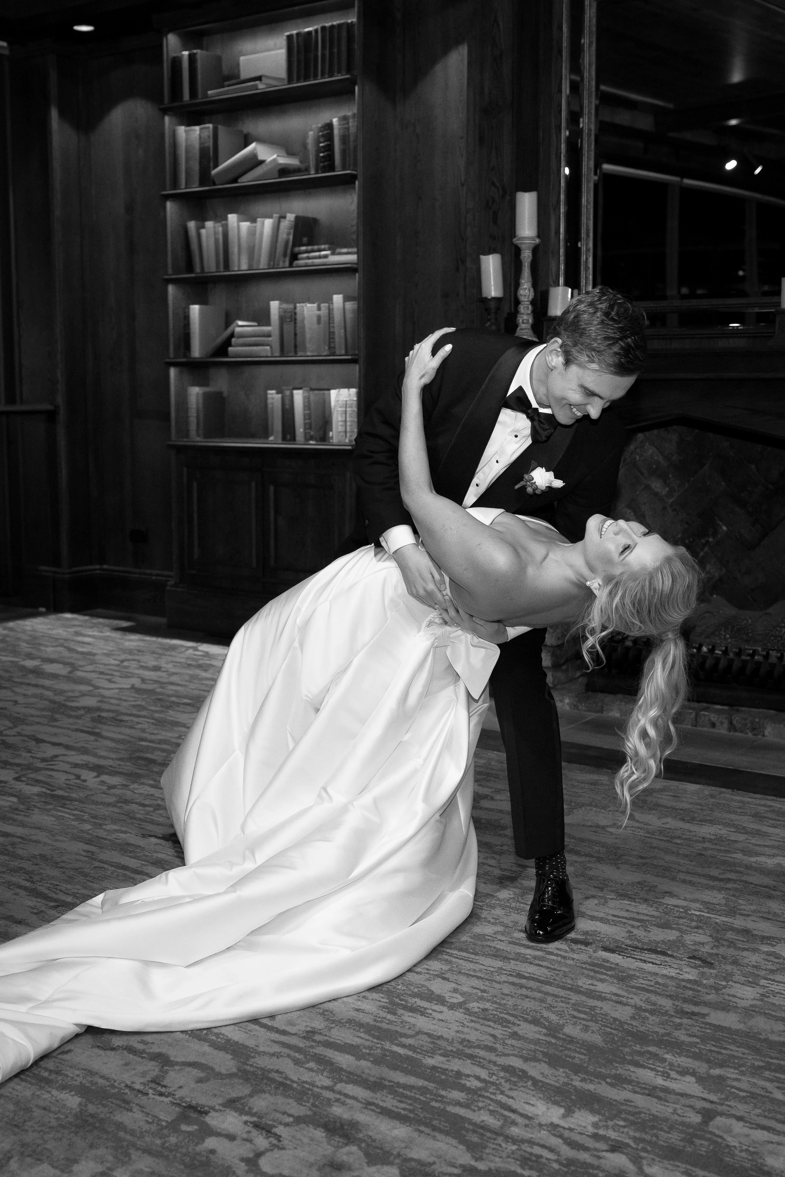 A black and white photo of a smiling groom dipping and kissing a bride in a wedding dress, in a cozy indoor setting with a bookshelf and fireplace in the background.