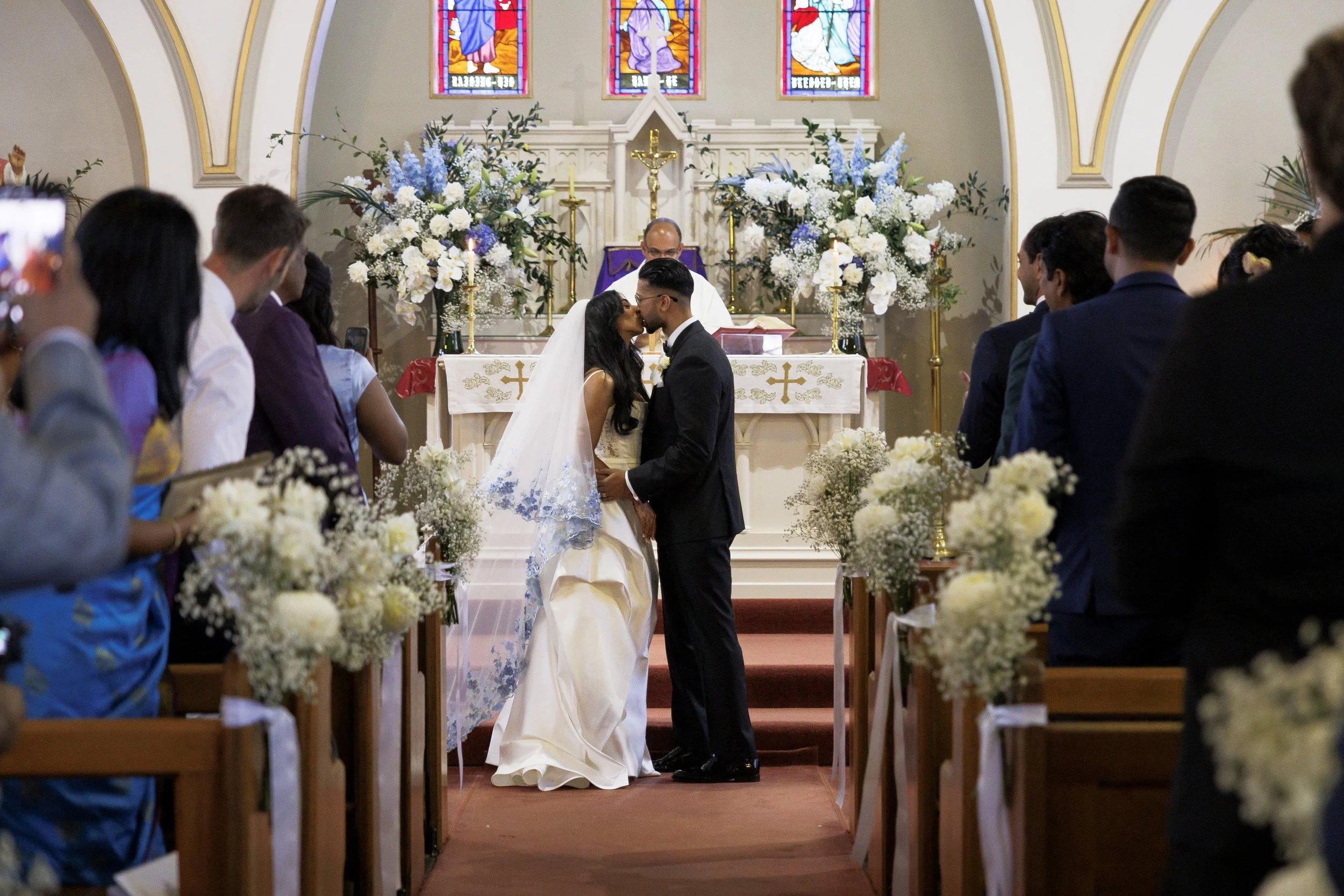 A wedding ceremony taking place inside a church with a bride and groom kissing at the altar, surrounded by floral arrangements and wedding guests.