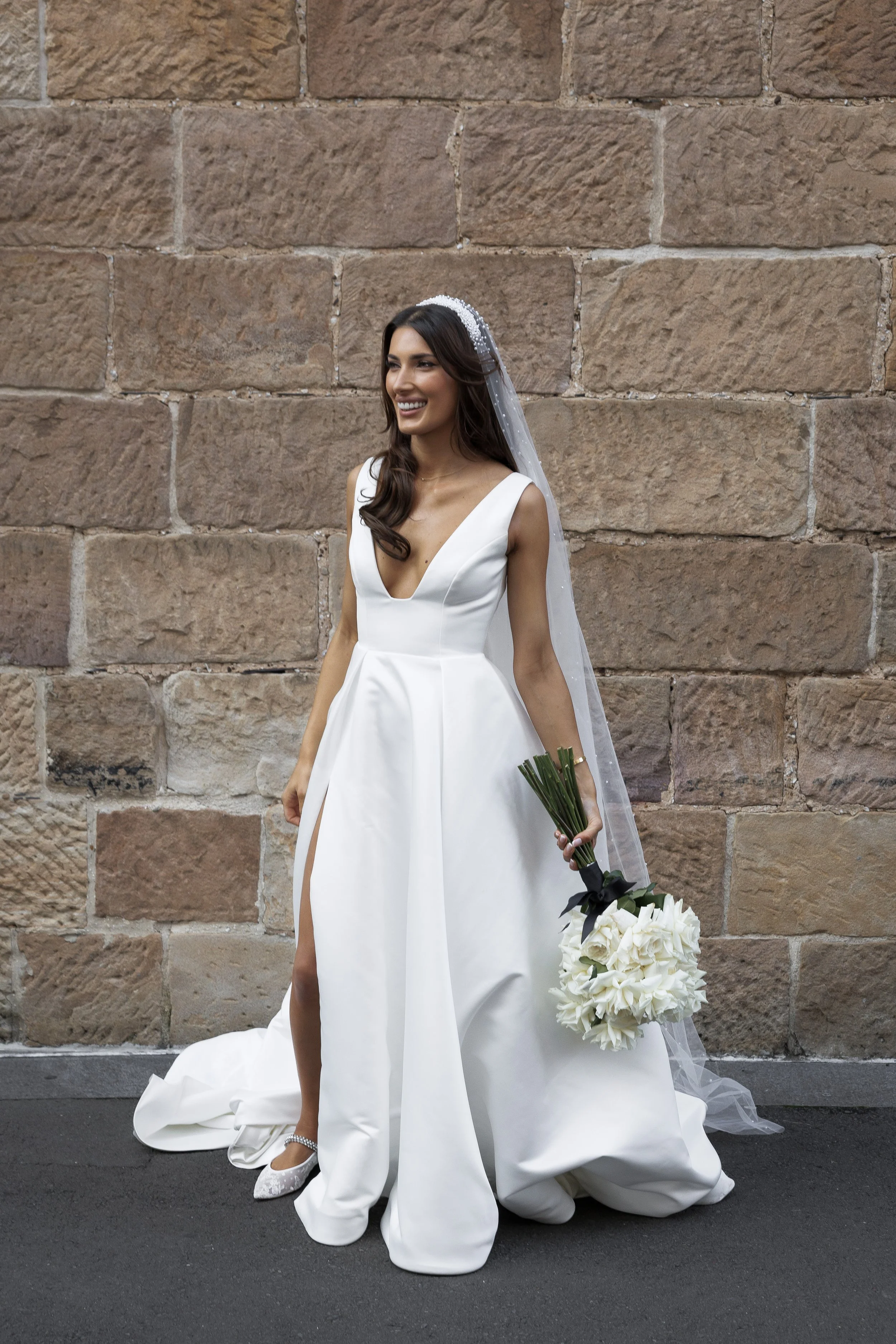 A bride in a white wedding gown smiling and holding a bouquet of white flowers, standing against a brick wall.