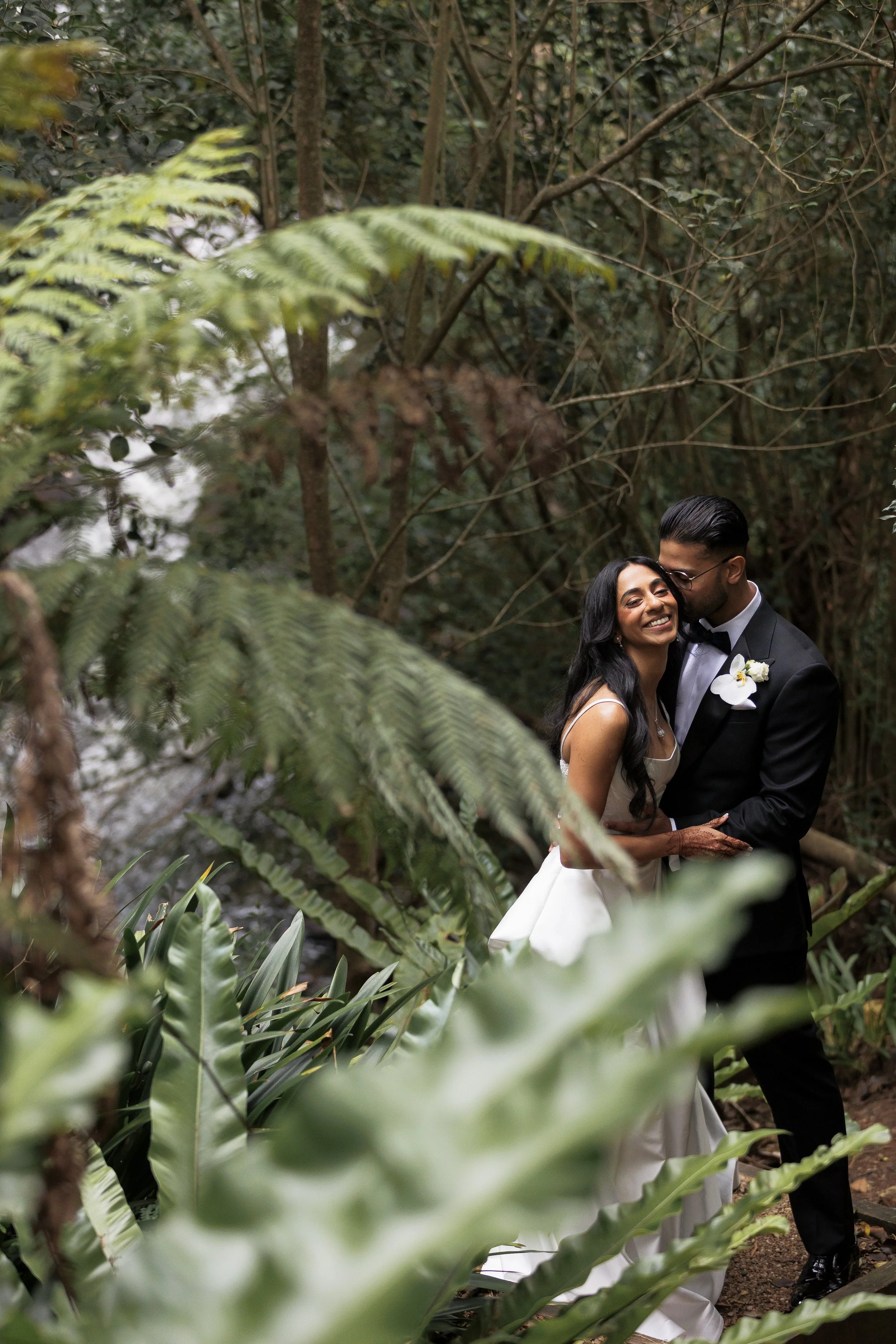 A bride and groom embracing and smiling in a lush, green forest, with large fern leaves and other foliage surrounding them.