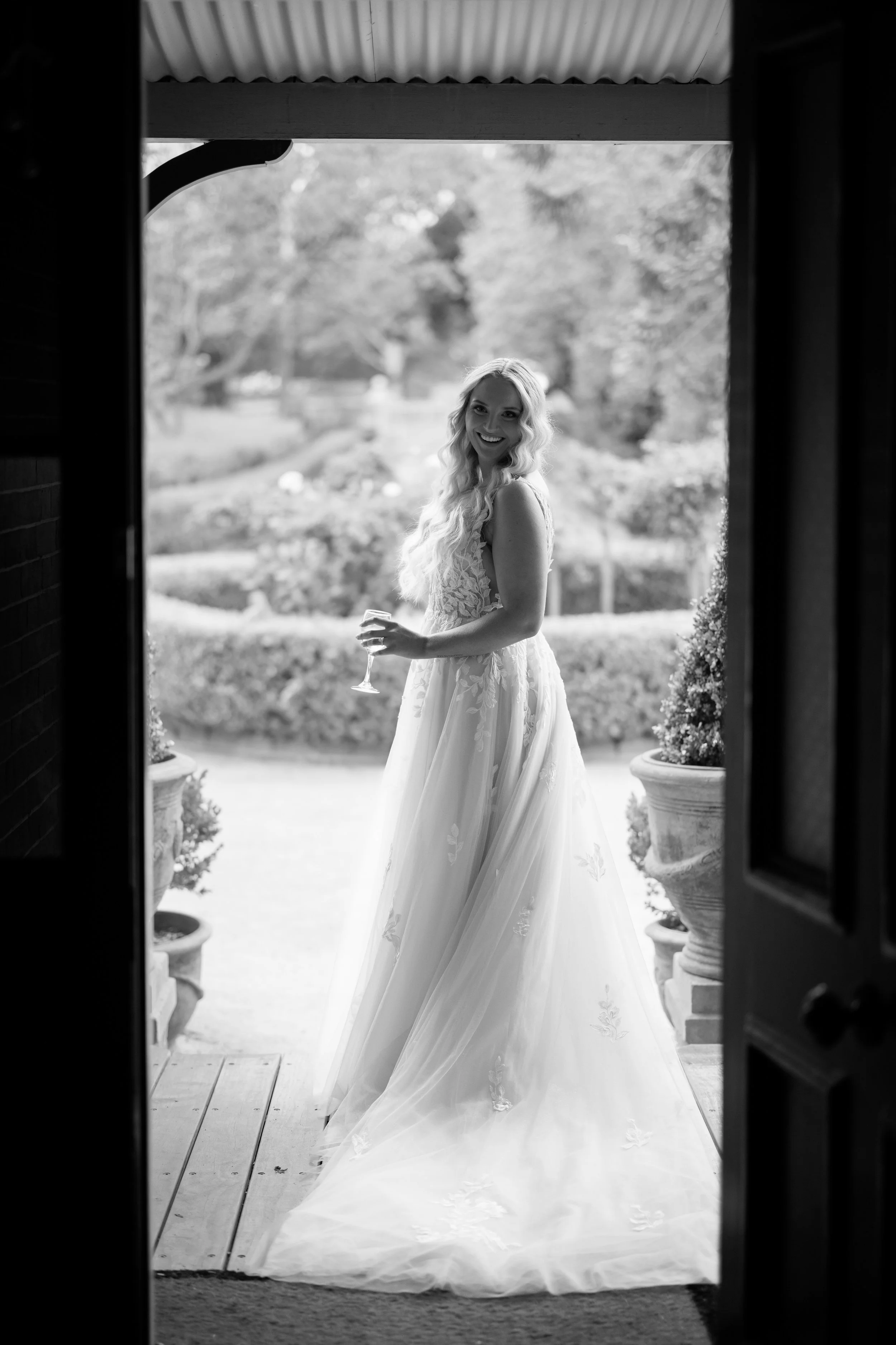 A woman in a wedding dress holding a glass of wine, smiling and standing outdoors on a porch with a garden in the background.