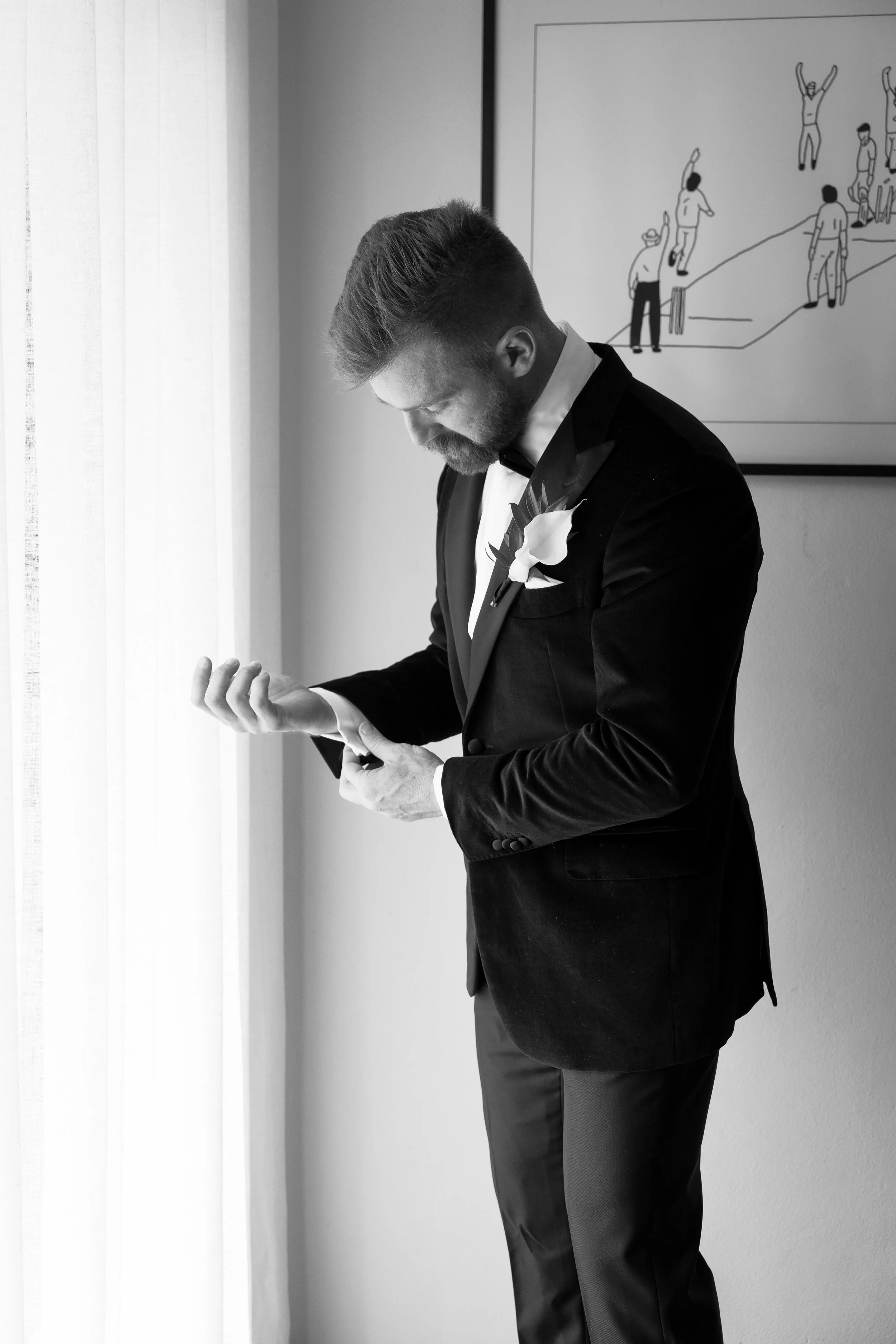 A man dressed in a tuxedo with a boutonniere is adjusting his sleeve near a window. There is a large framed drawing of people in various poses on the wall behind him.