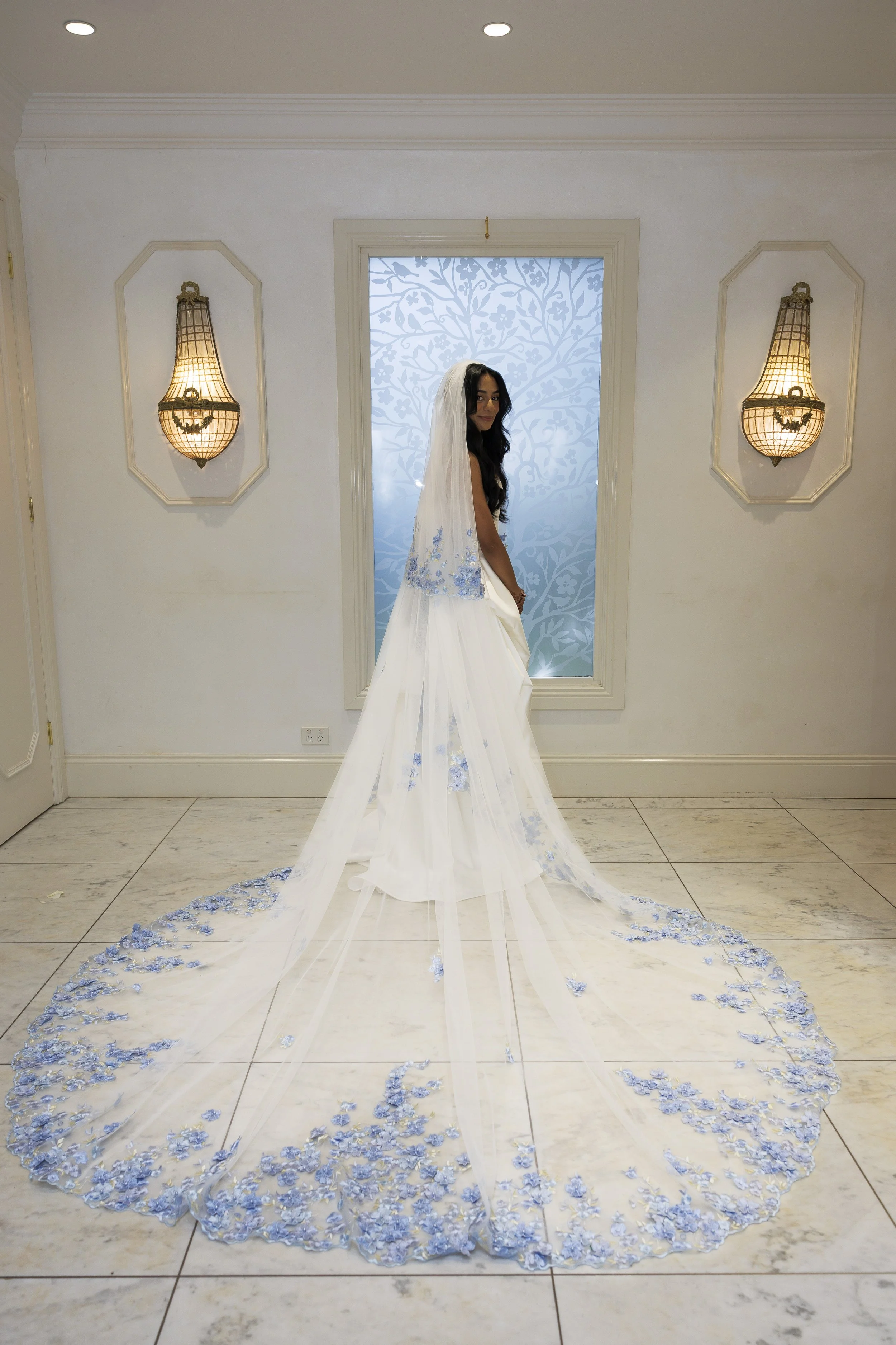 A woman in a white wedding dress with a blue floral pattern and floral lace train, standing indoors in front of a frosted window with a tree design, with two wall-mounted lamps on each side.