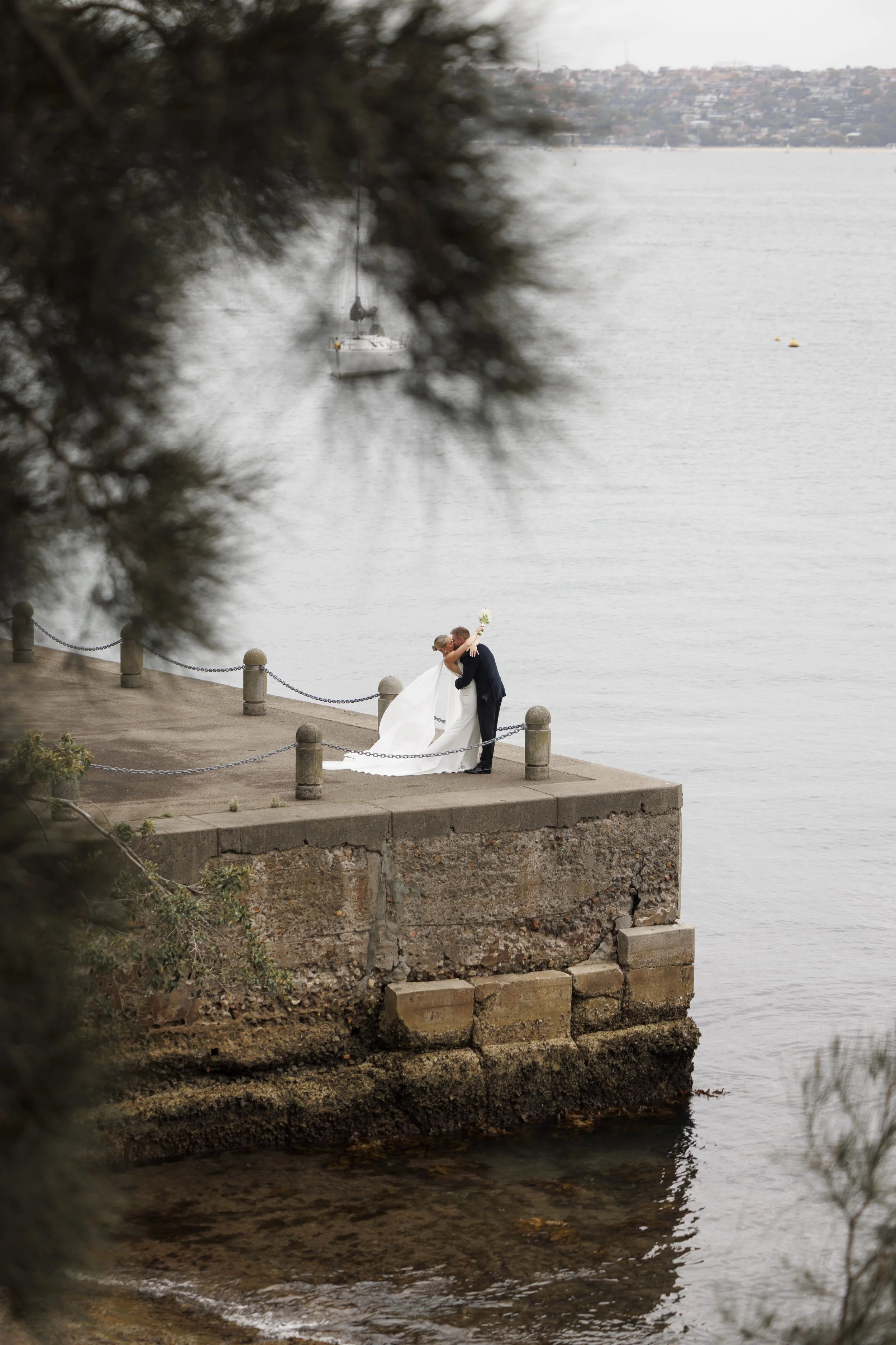 A newlywed couple sharing a kiss on a concrete pier by the water, with a sailboat in the background and trees framing the scene.