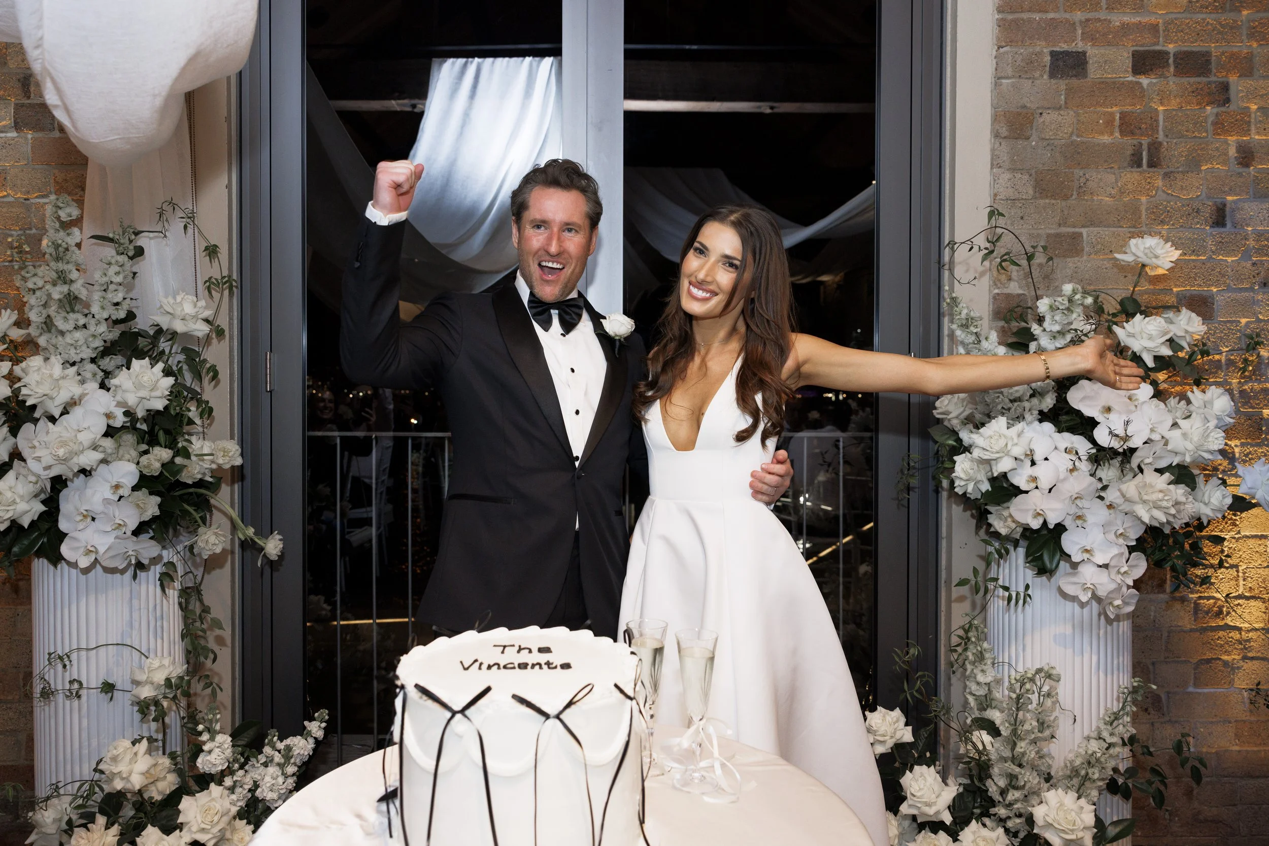 A happy bride and groom celebrating their wedding, standing with arms raised, in front of a table with a wedding cake and champagne glasses, decorated with white flowers.