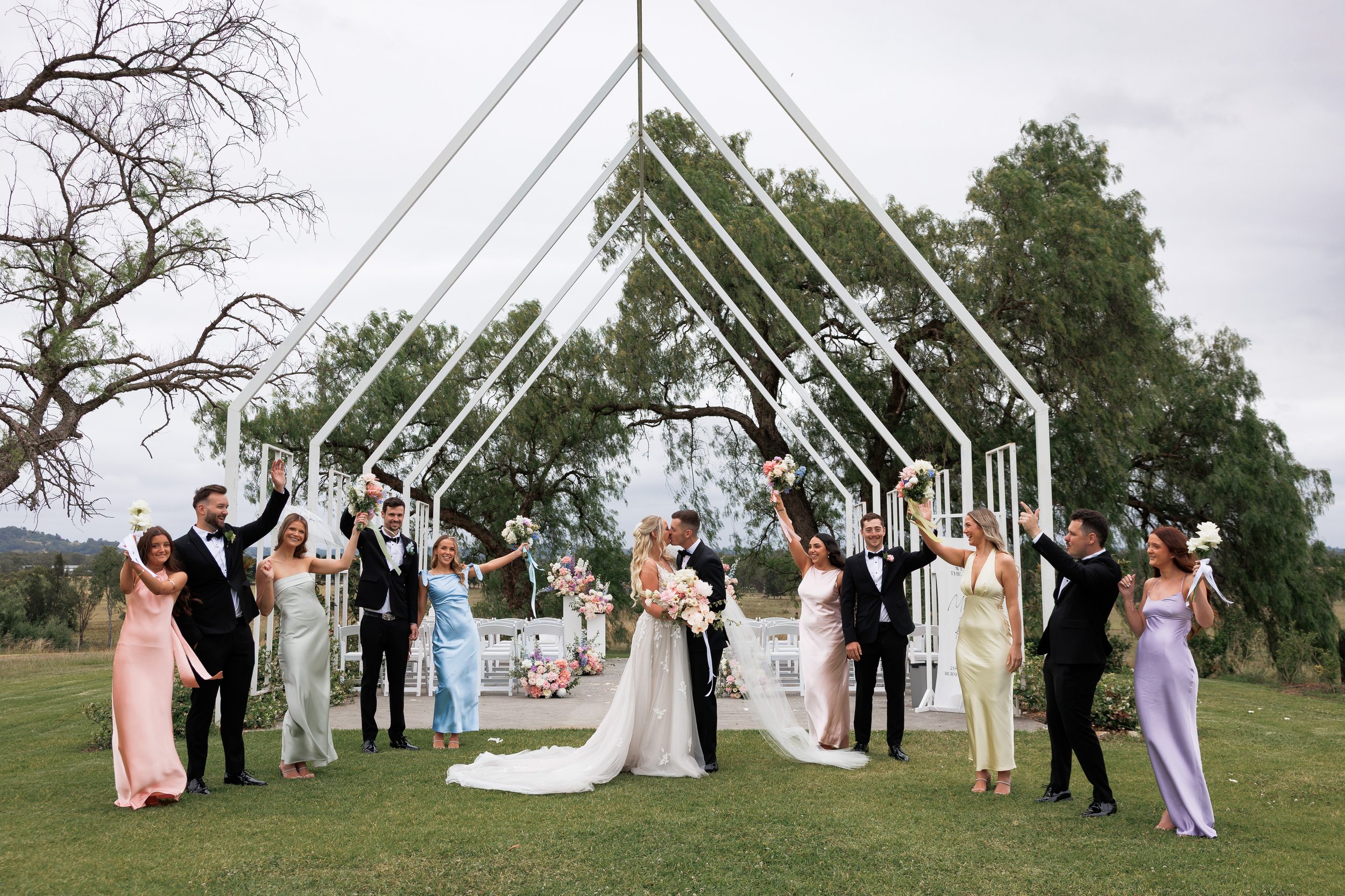 A wedding ceremony outdoors with the bride and groom kissing, surrounded by friends and family holding bouquets and raising their hands in celebration, under a white canopy frame with trees and a cloudy sky in the background.
