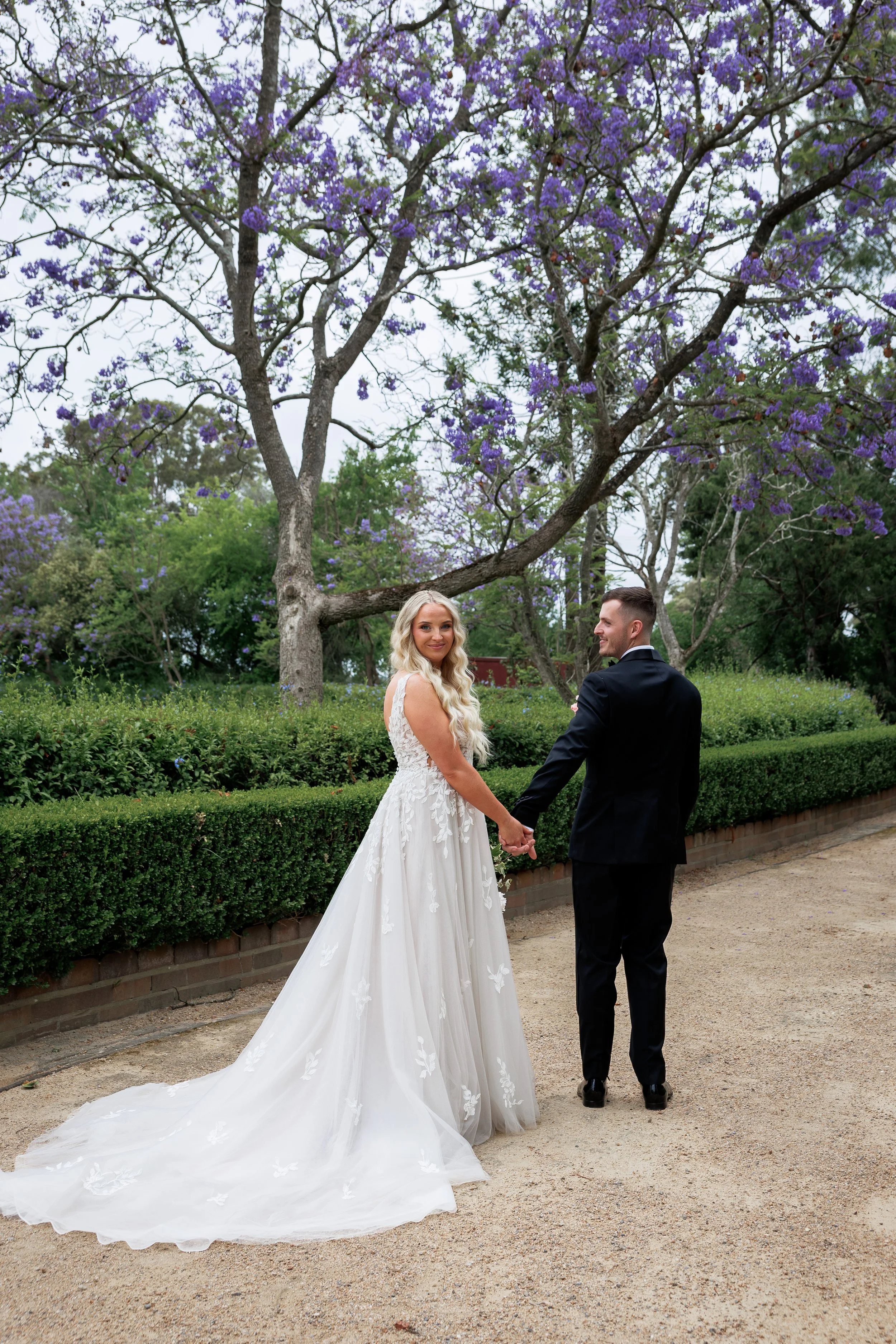 A bride and groom holding hands, standing outdoors on a dirt pathway, with purple flowering trees and green bushes in the background.