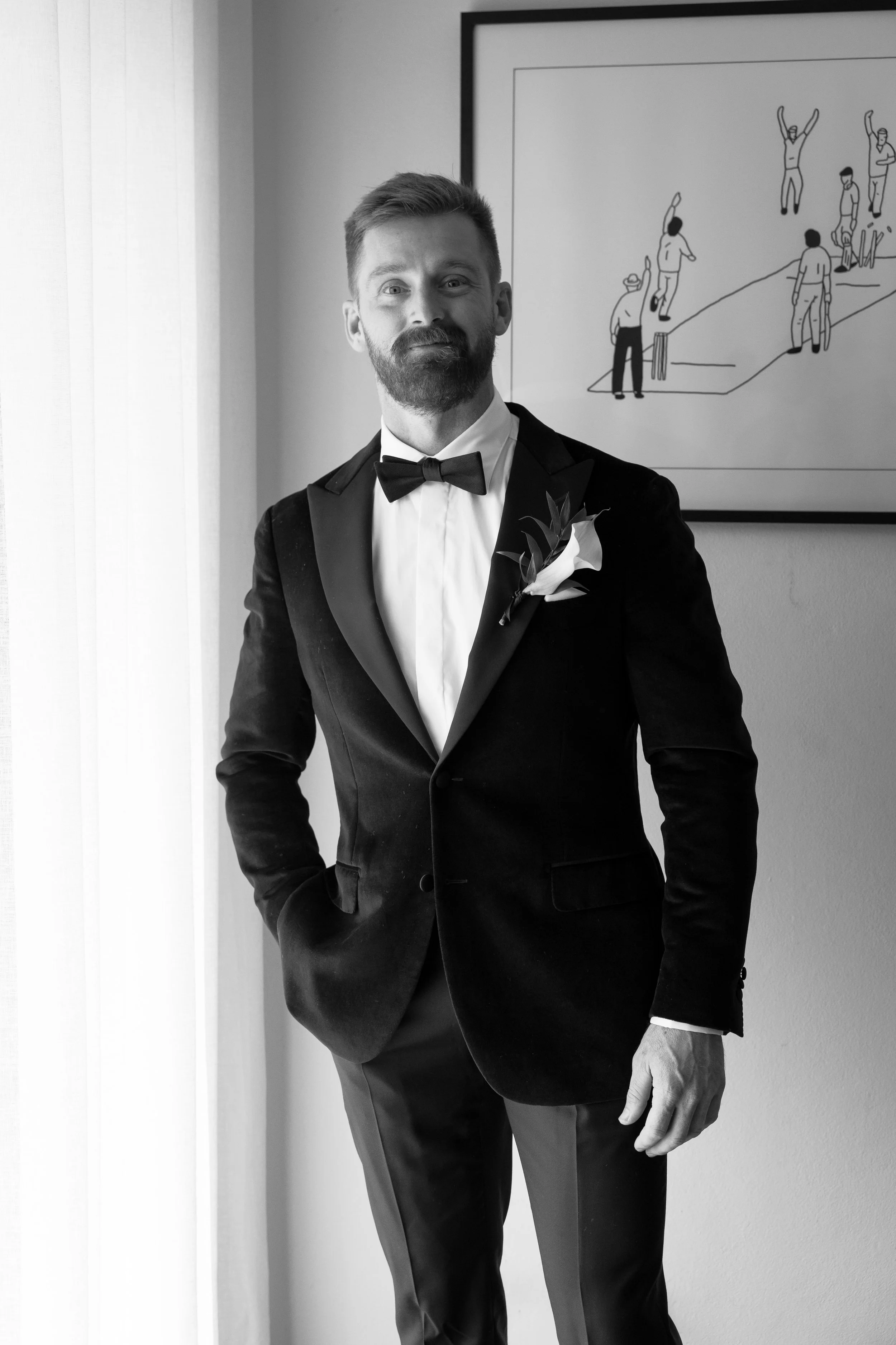 Black and white photo of a man in a tuxedo with a bow tie and a white flower boutonniere, standing near a window next to a wall with a framed drawing of people playing cricket.