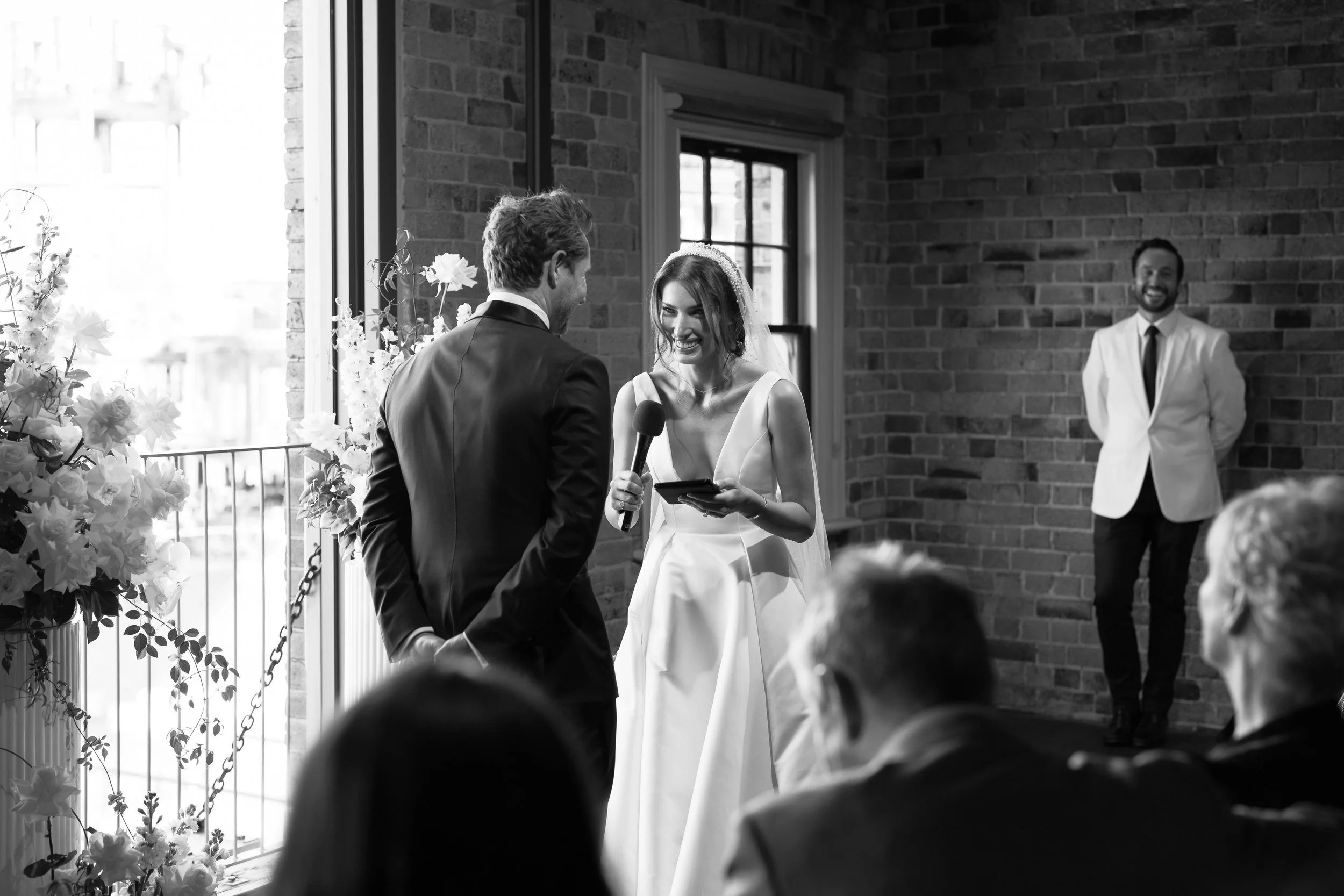 A bride and groom exchanging vows during a wedding ceremony indoors, with the bride holding a microphone and reading from a tablet, while a man in a white blazer stands in the background smiling.