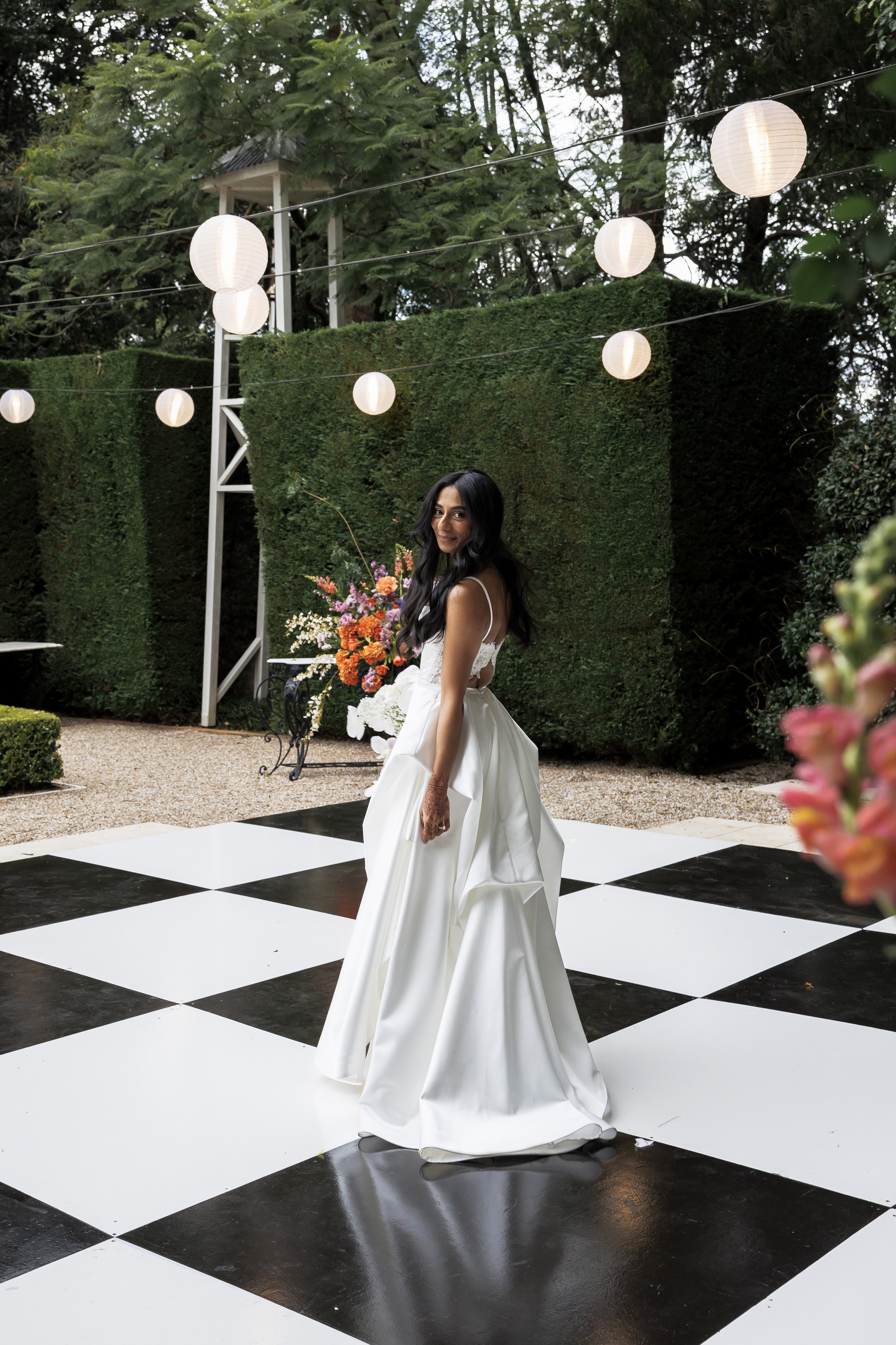 A woman in a white dress standing on a black and white checkered dance floor at an outdoor event, surrounded by greenery and hanging white lanterns.