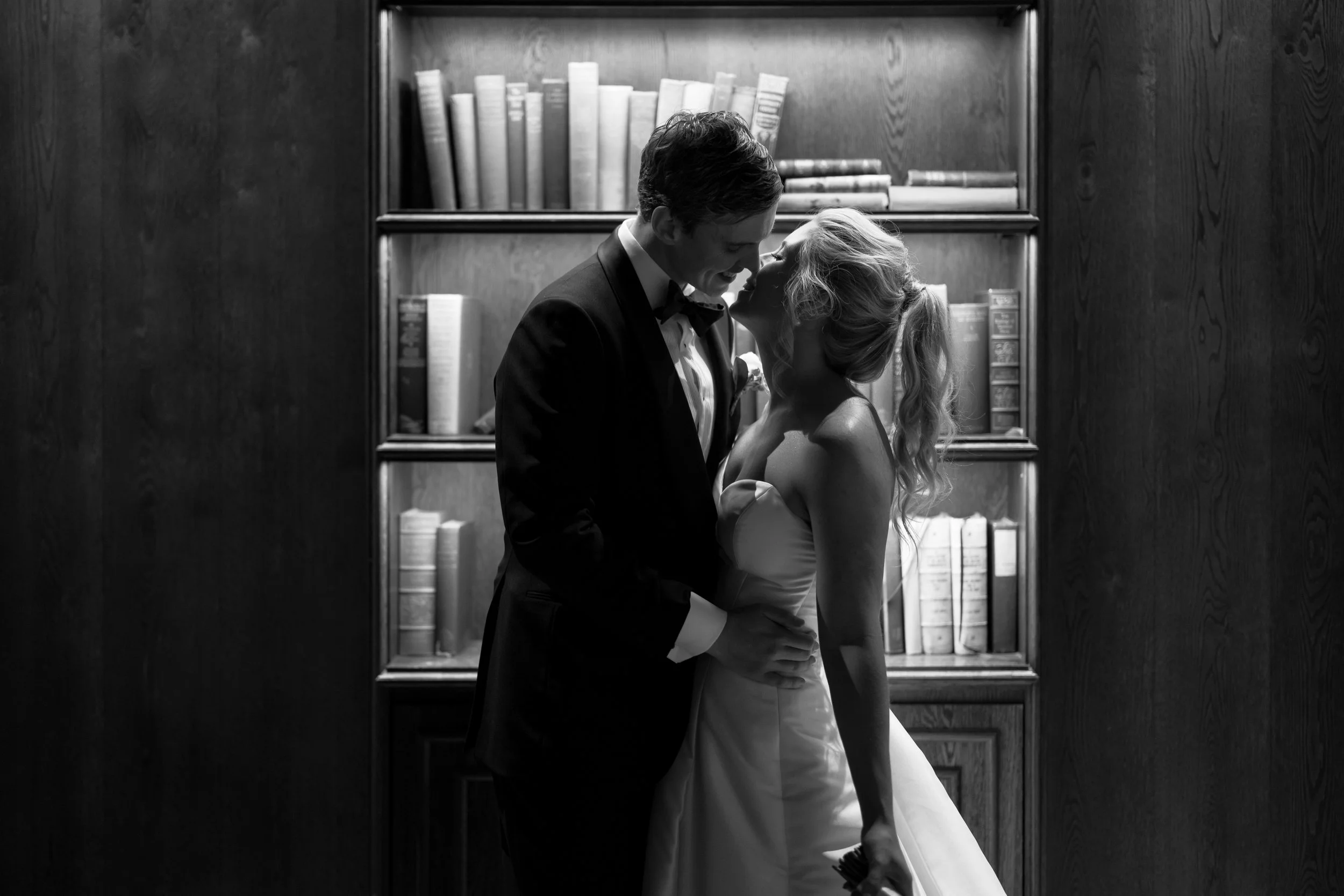 A black and white photo of a bride and groom in wedding attire sharing a kiss in front of a bookshelf filled with books.