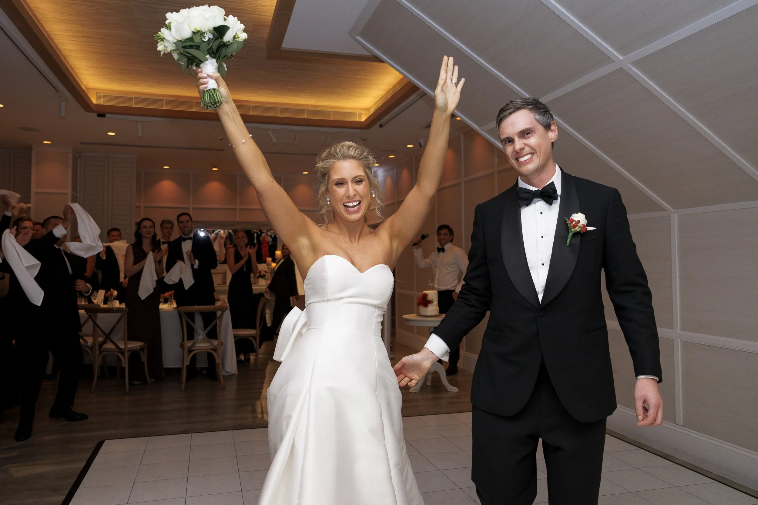 Bride and groom celebrating at their wedding reception, with the bride holding a bouquet of white flowers and raising her arms, while the groom holds her hand, all smiling.