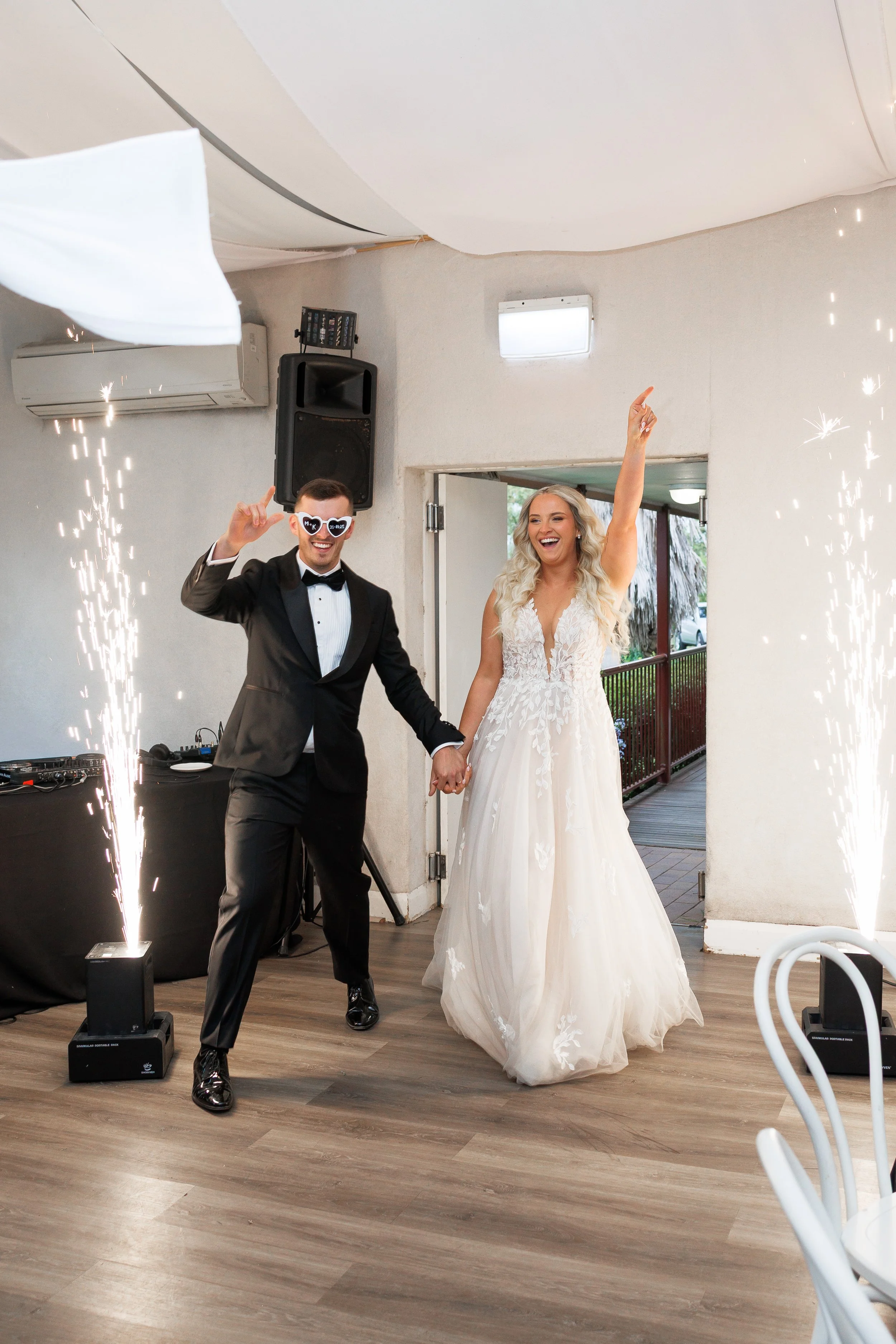 A bride and groom celebrating at their wedding reception with sparklers, holding hands, the groom is wearing a tuxedo and funny glasses, and the bride is wearing a white wedding dress and smiling with her arm raised.