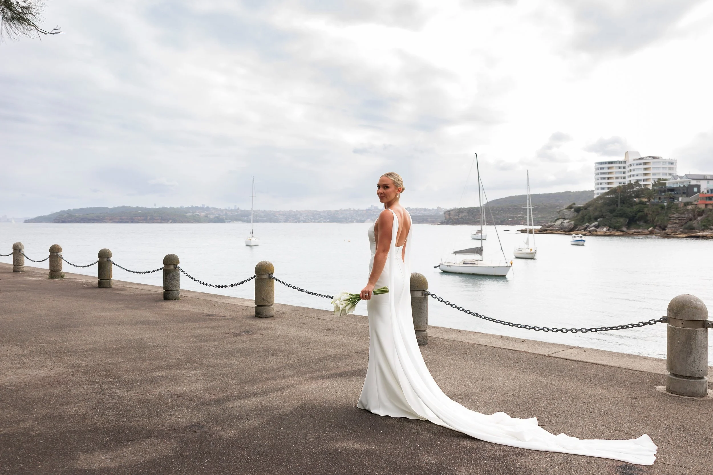 A woman in a white wedding dress holding a bouquet of flowers standing on a waterfront promenade with boats and sailboats on the water, and buildings on a hill in the background.
