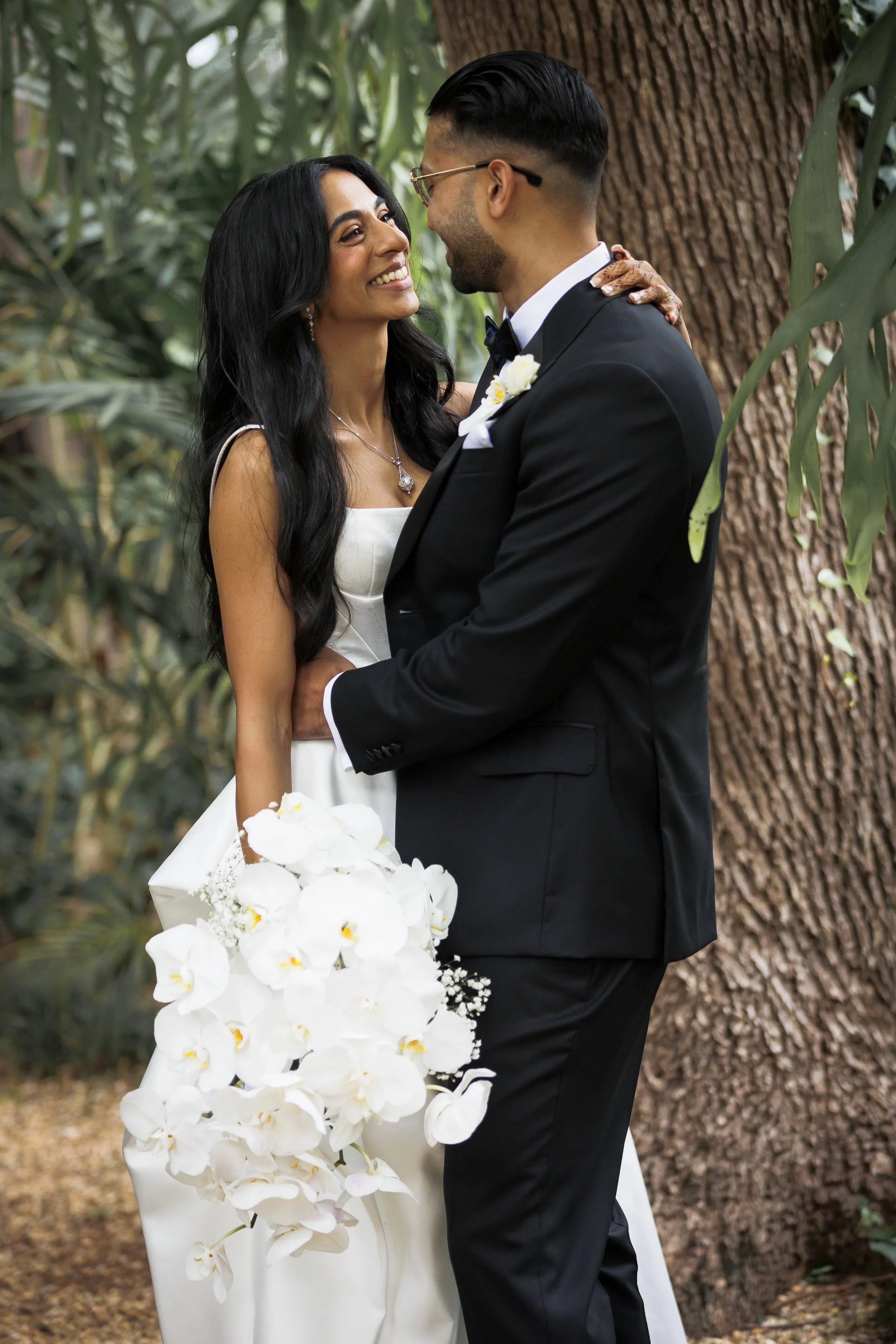 A bride and groom smile at each other, embracing in a lush, green outdoor setting with a large tree trunk in the background. The bride holds a cascading white orchid bouquet, and the groom wears a black tuxedo with a bow tie and white boutonniere.