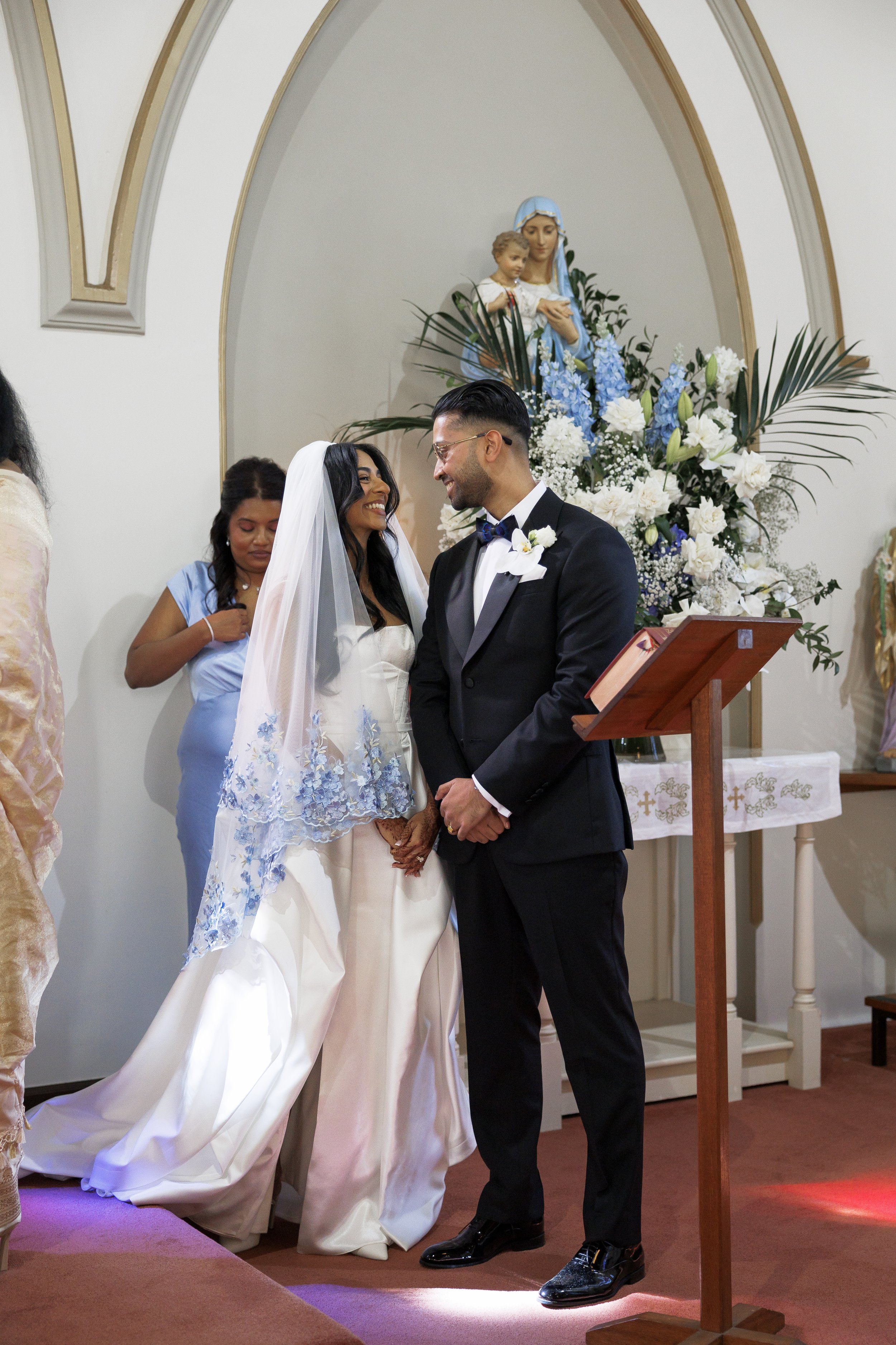 A bride and groom stand facing each other, holding hands, during a wedding ceremony inside a church with religious decor and a biblical statue in the background.