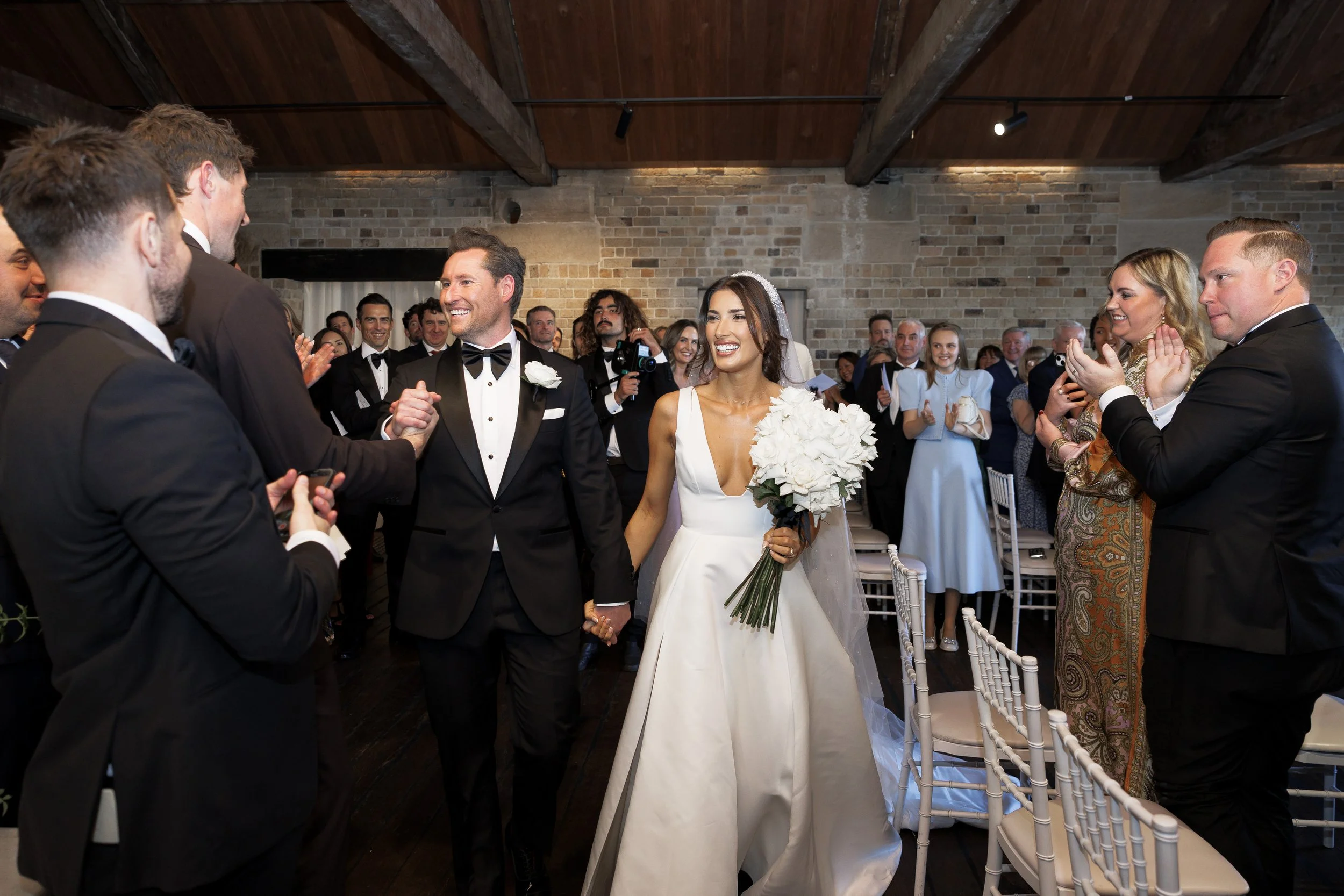 Bride and groom walking down aisle, holding hands, smiling surrounded by wedding guests clapping and celebrating indoors with brick walls and wooden ceiling beams.