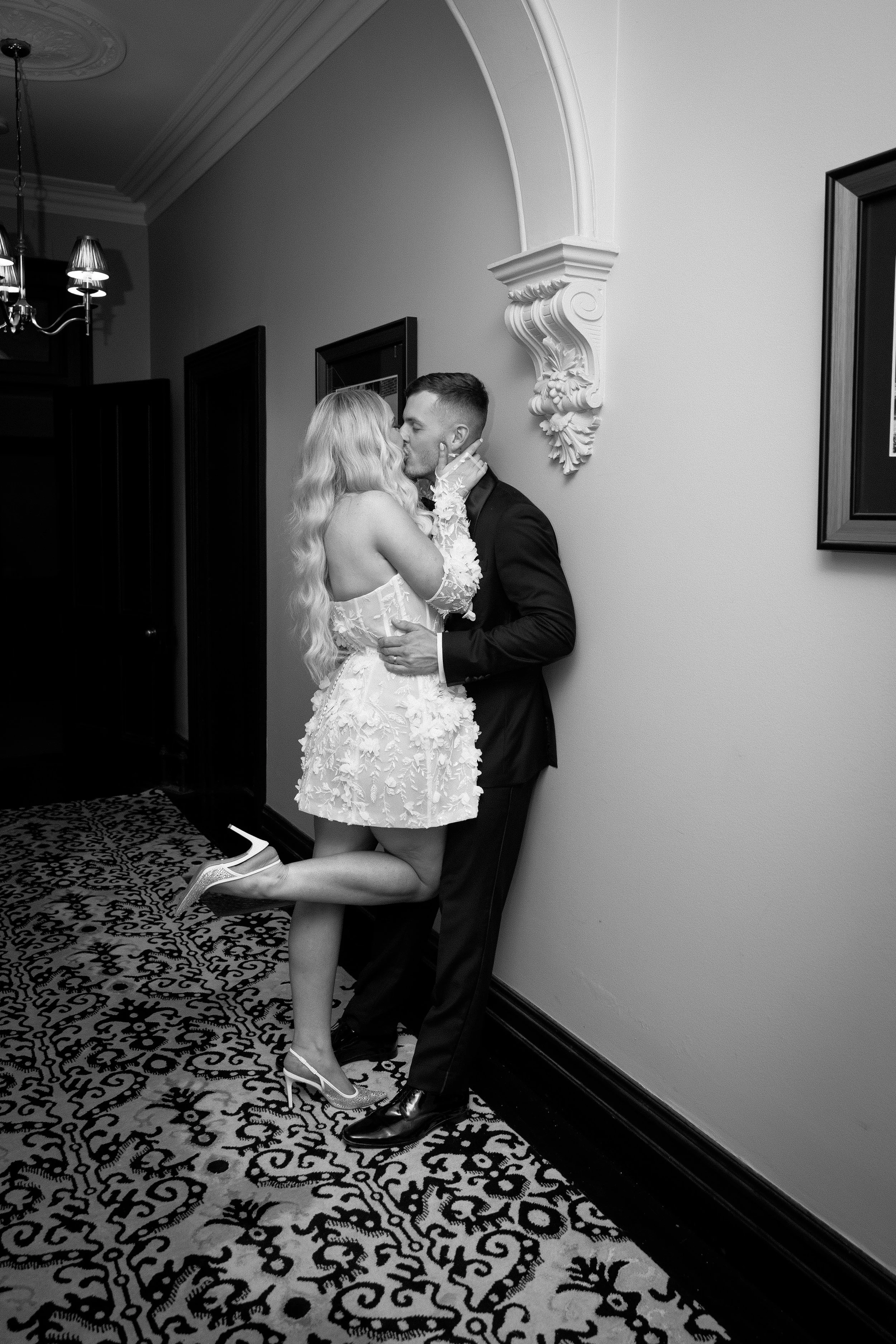 A couple sharing a romantic kiss indoors, the woman wearing a detailed white dress and heels, the man in a dark suit, with ornate wall decor and patterned carpet visible.