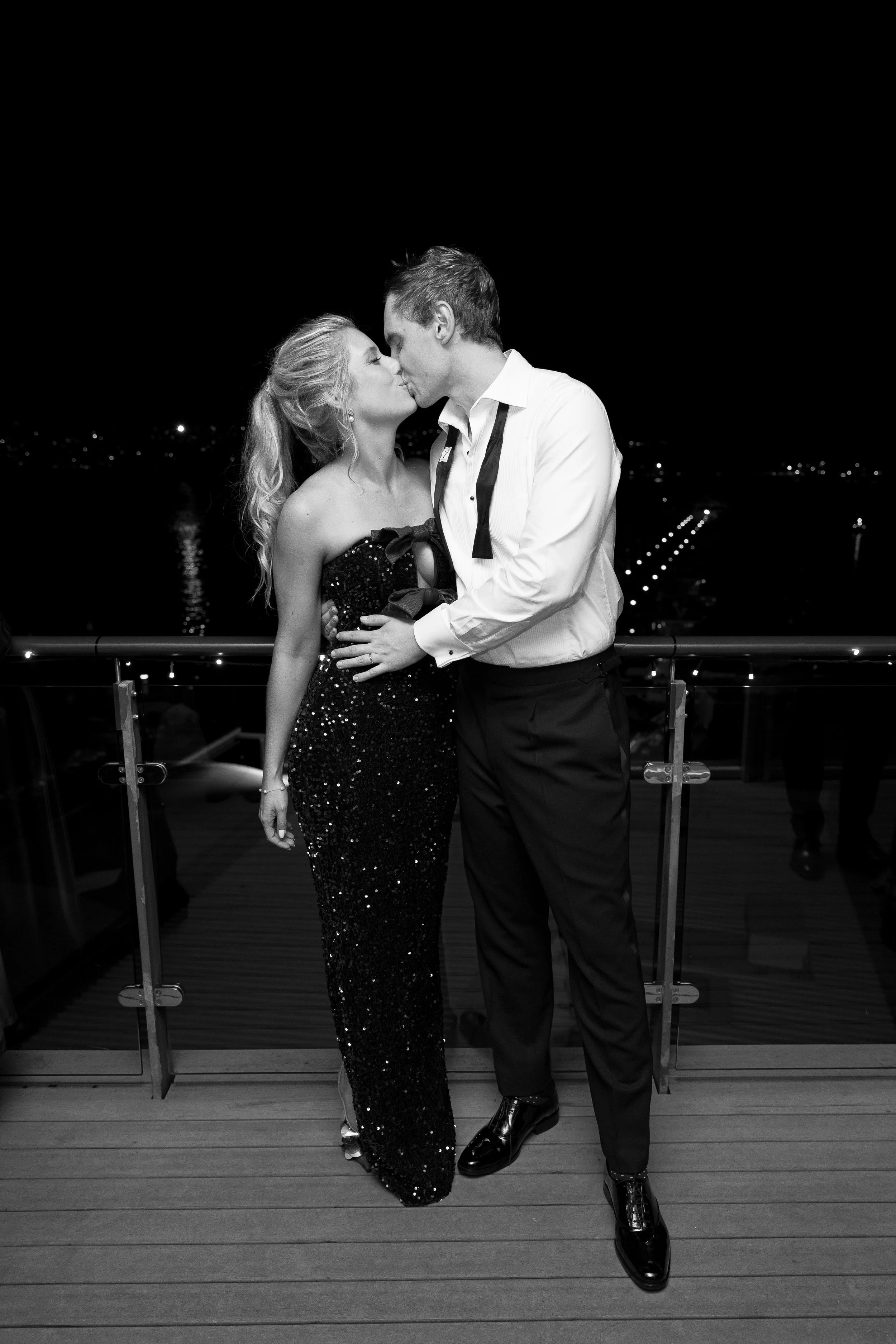 A couple in formal attire sharing a kiss at night on a balcony overlooking a body of water and city lights.