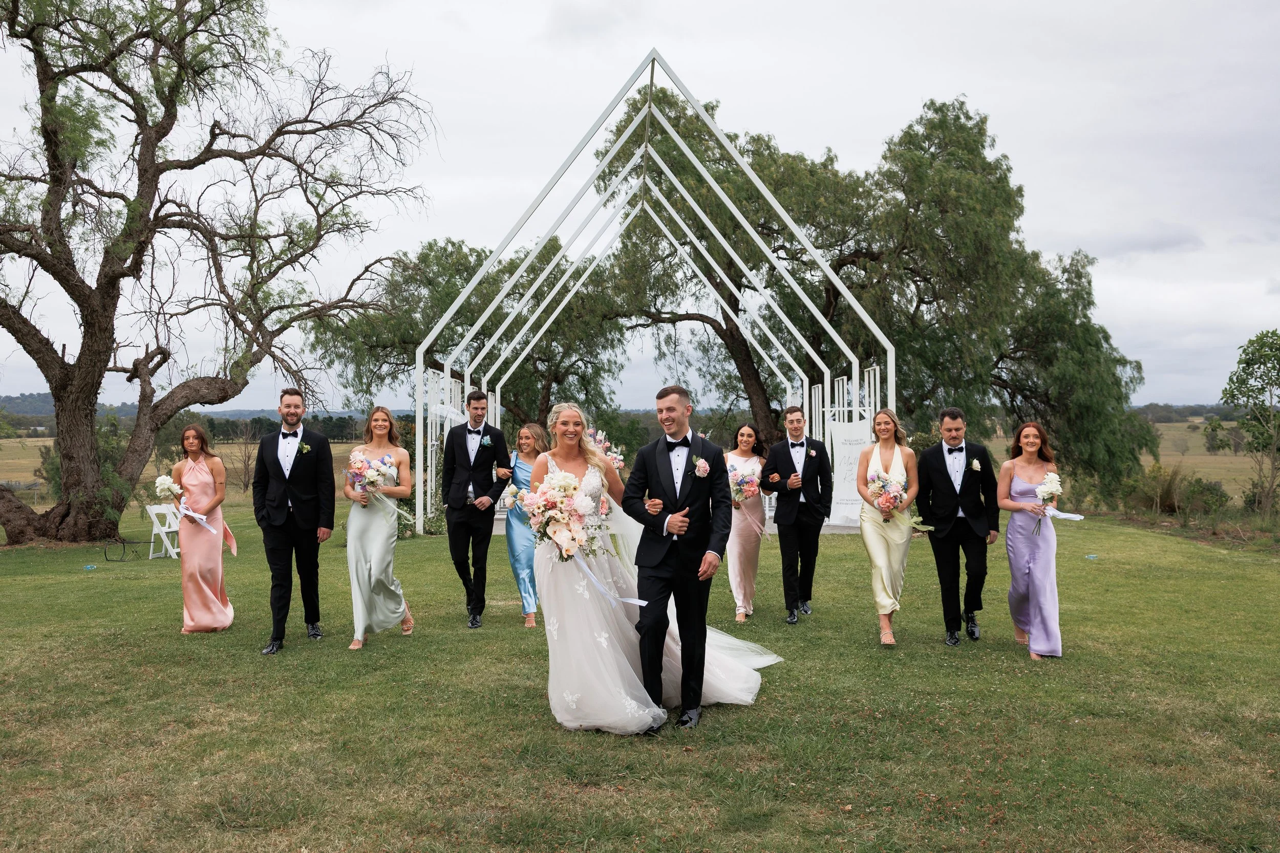 A wedding party walking on a grassy field outdoors. The bride and groom are in front, smiling and holding hands, with the bride holding a bouquet of flowers. Behind them are bridesmaids in pastel dresses and groomsmen in tuxedos. In the background, t