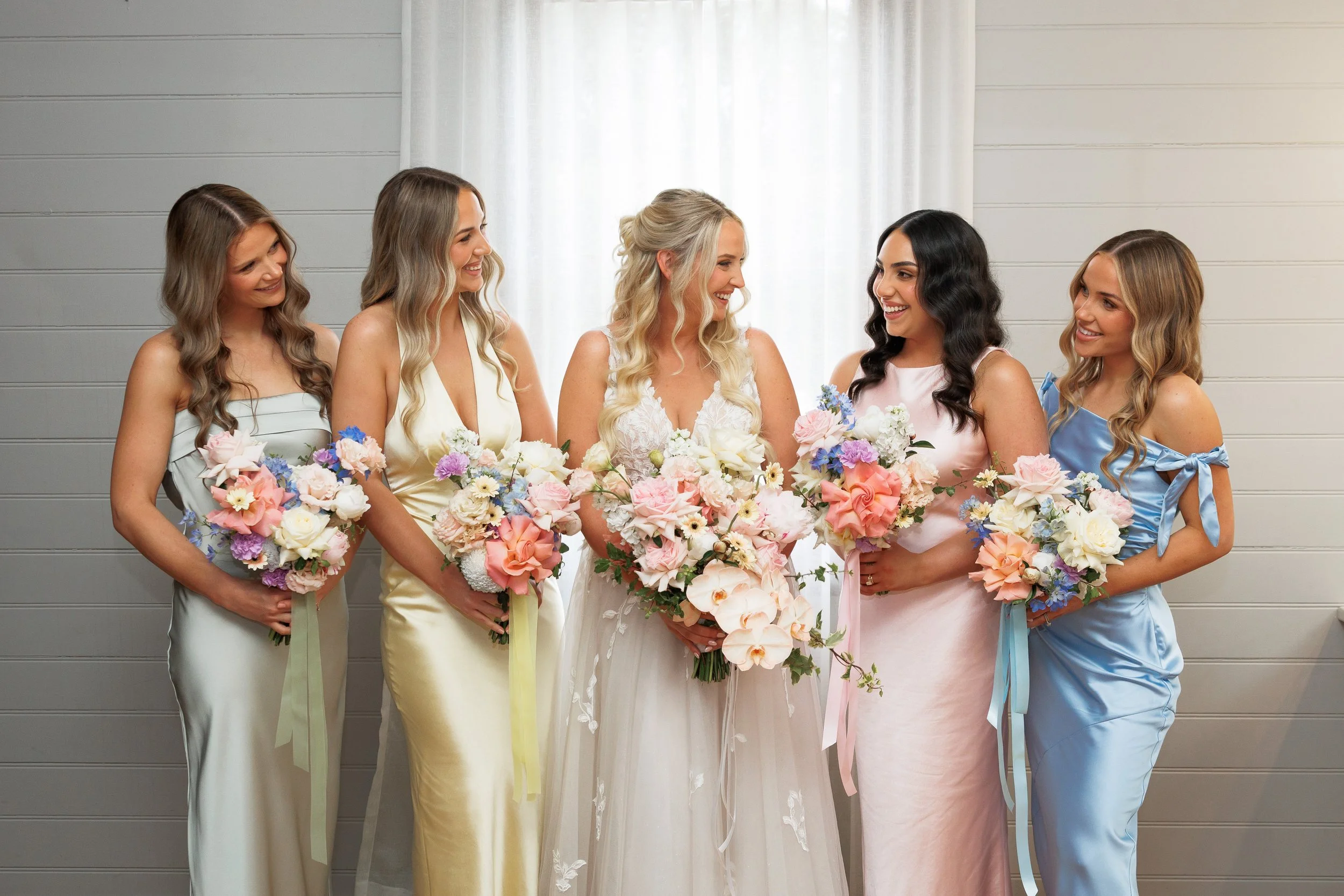 Bride and four bridesmaids at a wedding, holding bouquets of flowers, standing against a light gray wall with white curtains.