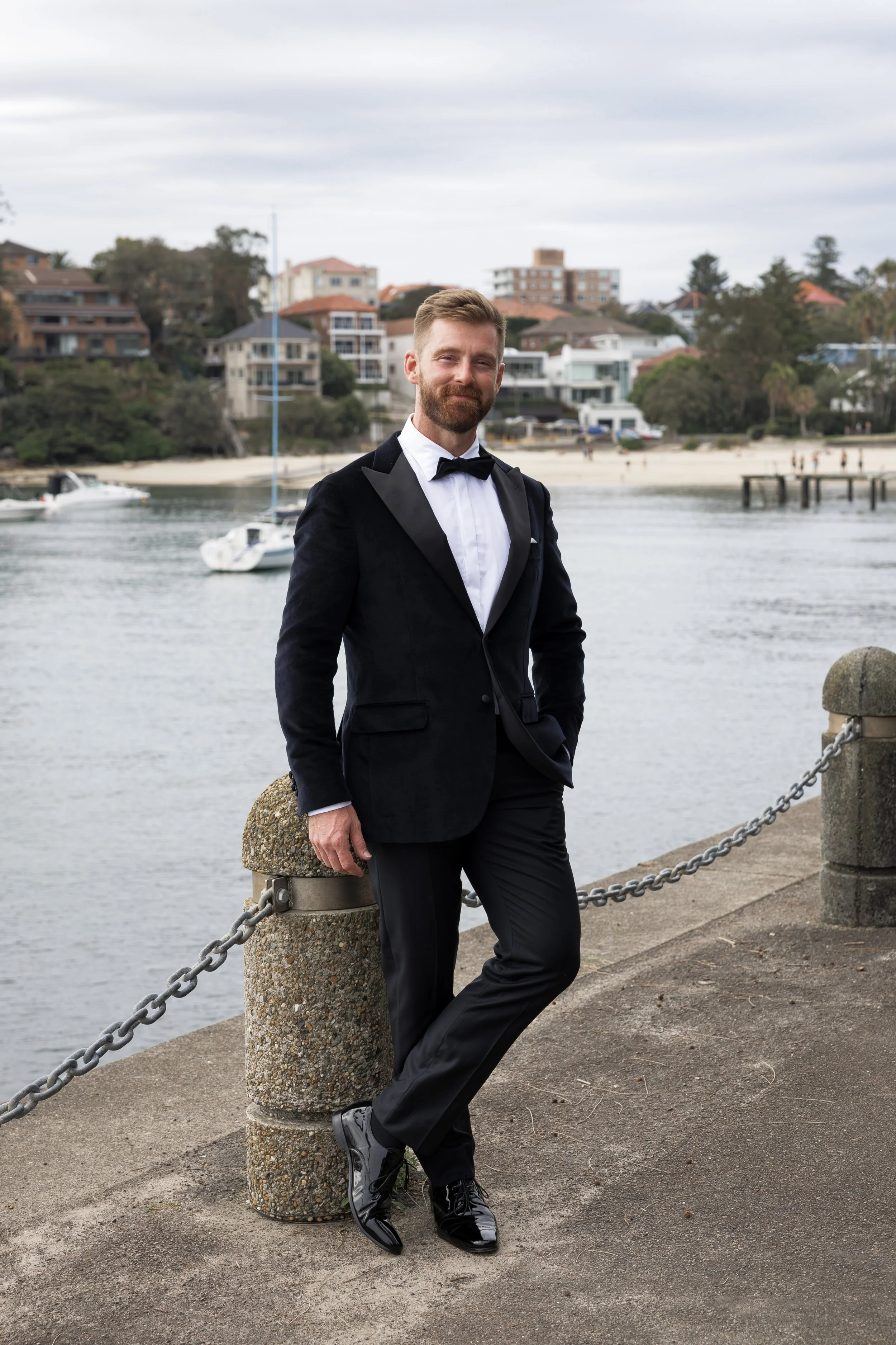 A man in a black tuxedo with a white shirt and black bow tie, standing on a waterfront promenade with a chain barrier, with boats and houses on a hill in the background.