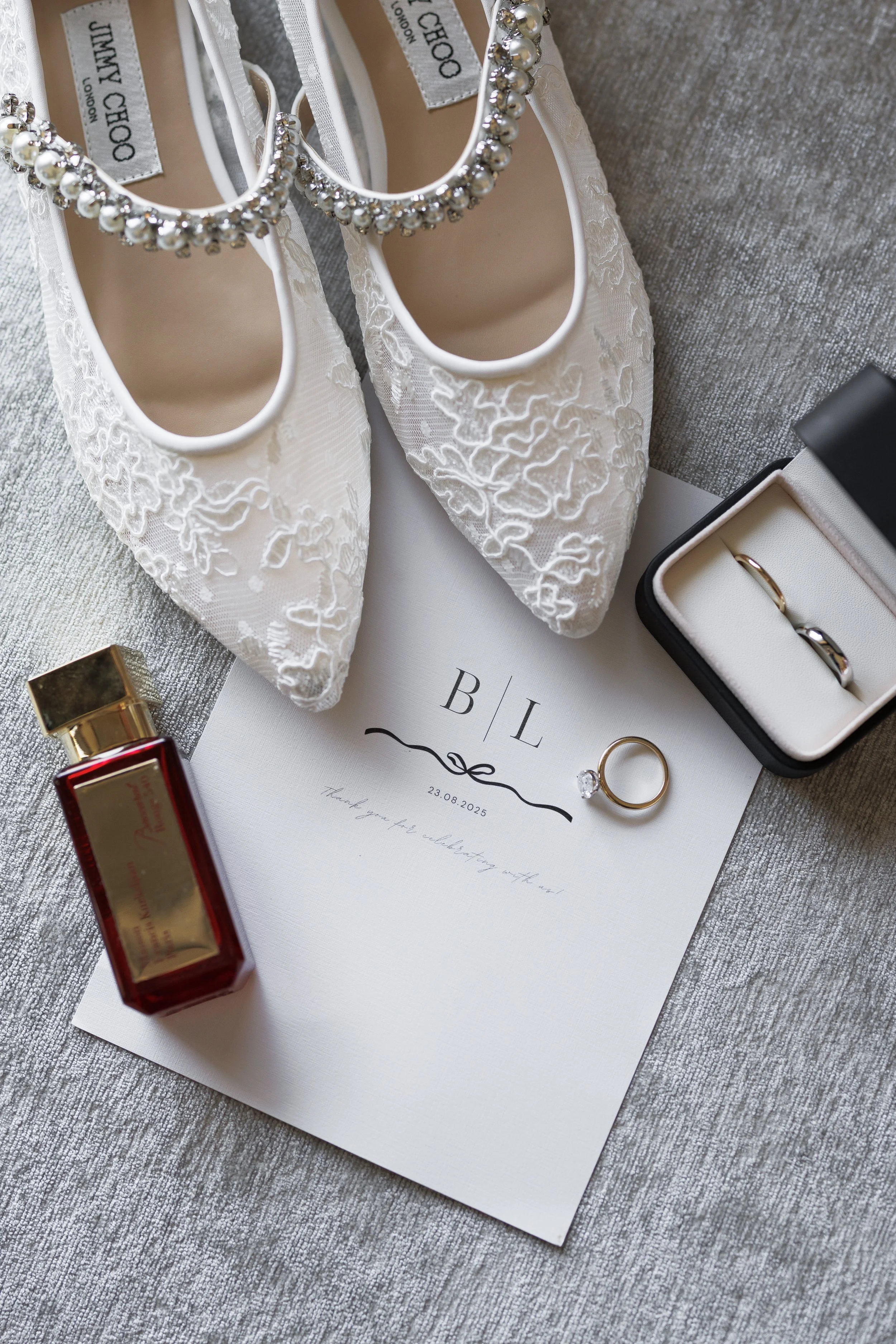 White lace wedding shoes with pearl and rhinestone straps, a wedding invitation with initials 'B' and 'L' dated 23.08.2025, a pair of wedding rings in a black and white ring box, a small bottle of perfume, and a wedding thank you note, all arranged o