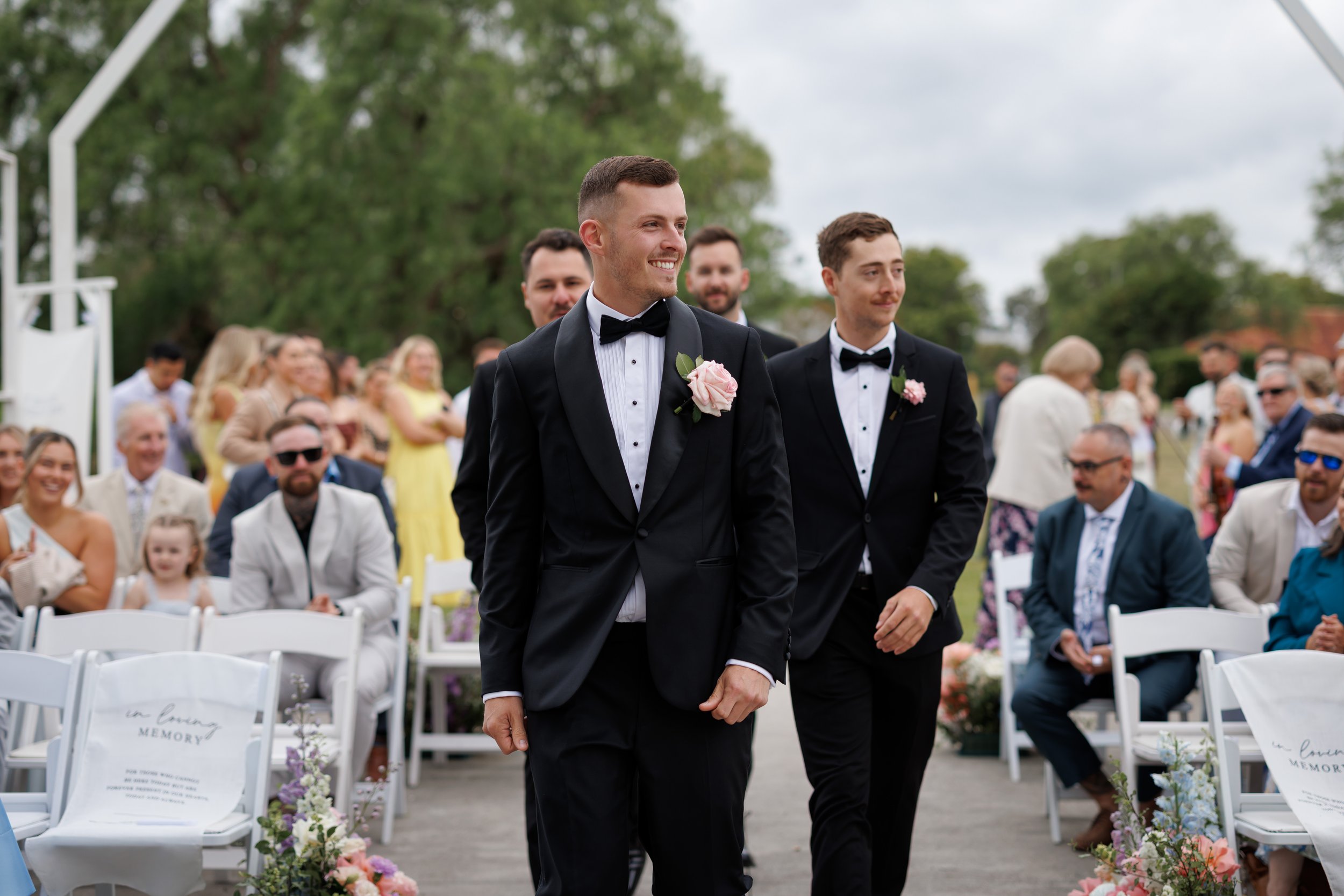 Groom and groomsman walking down aisle at outdoor wedding ceremony, guests seated on either side, greenery and cloudy sky in background.