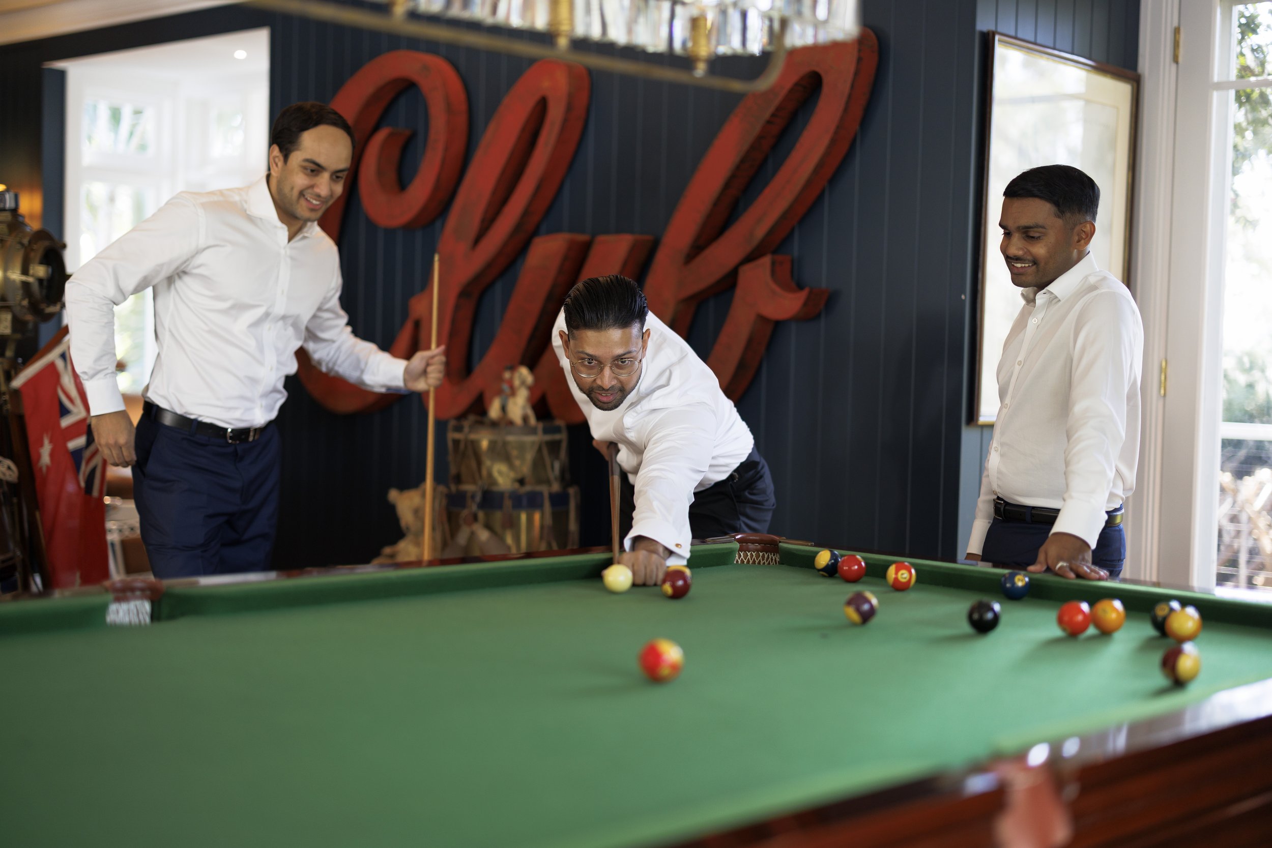 Three men playing billiards in a room with a large red 'Yard' sign on the wall, with one man aiming to hit the balls on the pool table while the other two watch.