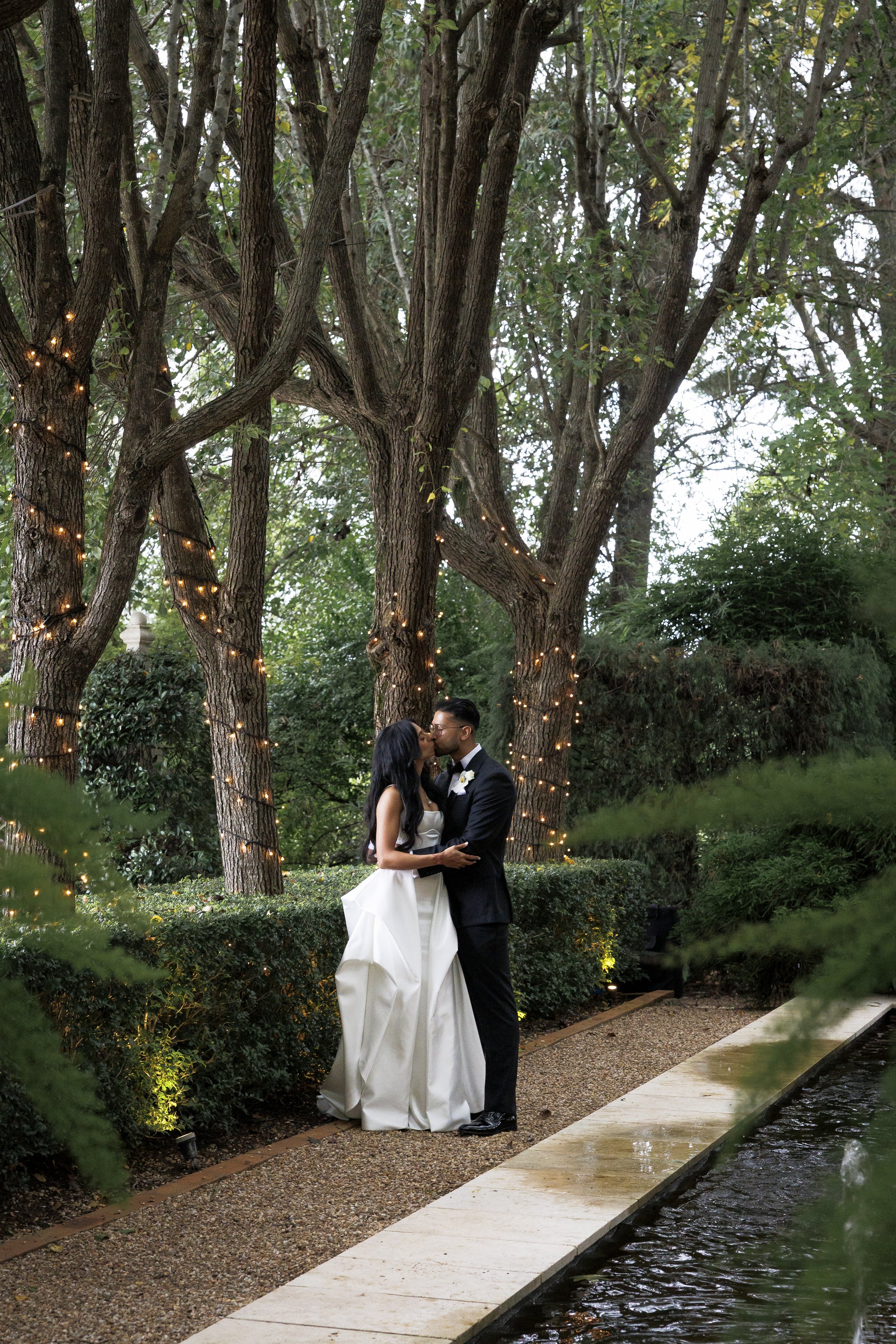 A bride and groom kissing outdoors on a wedding day, standing on a gravel pathway next to a water feature, surrounded by trees with string lights in the background.