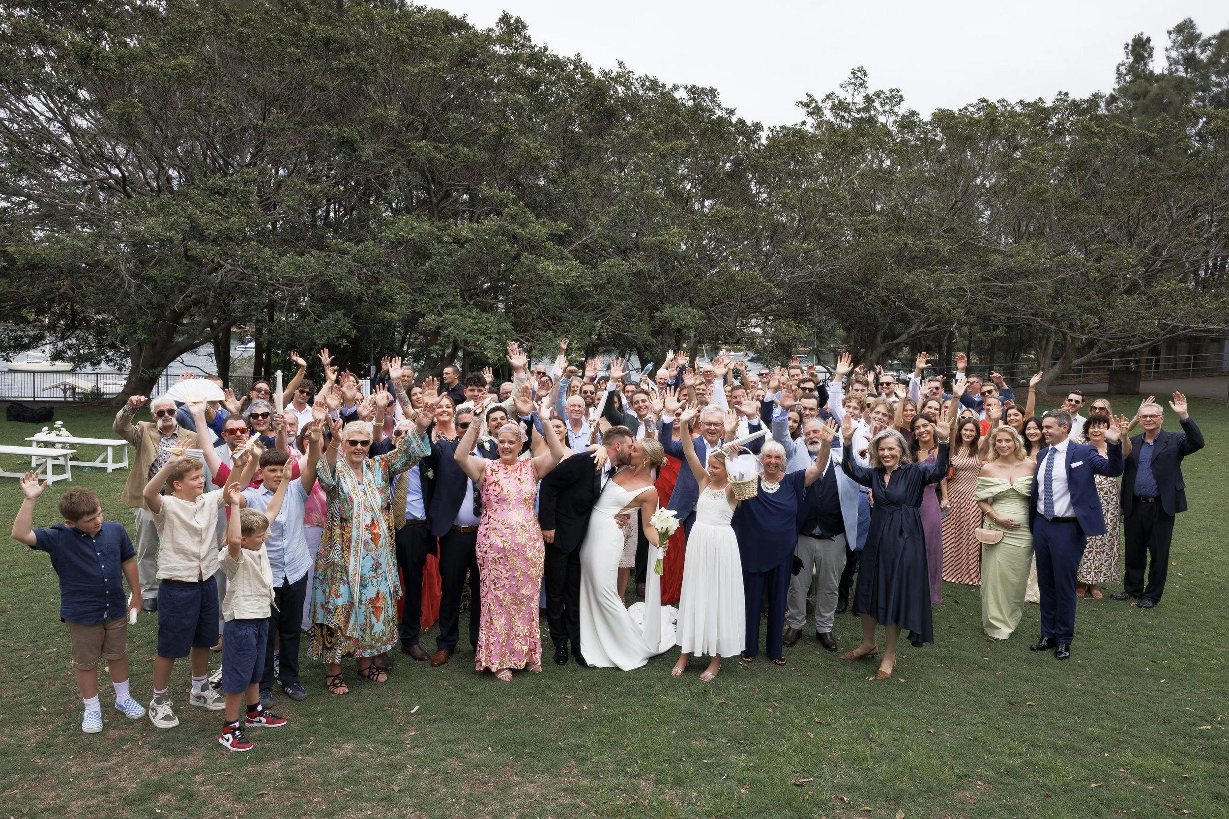 Group of wedding guests outdoors, cheering with hands raised, some dressed in formal attire, with trees in the background.