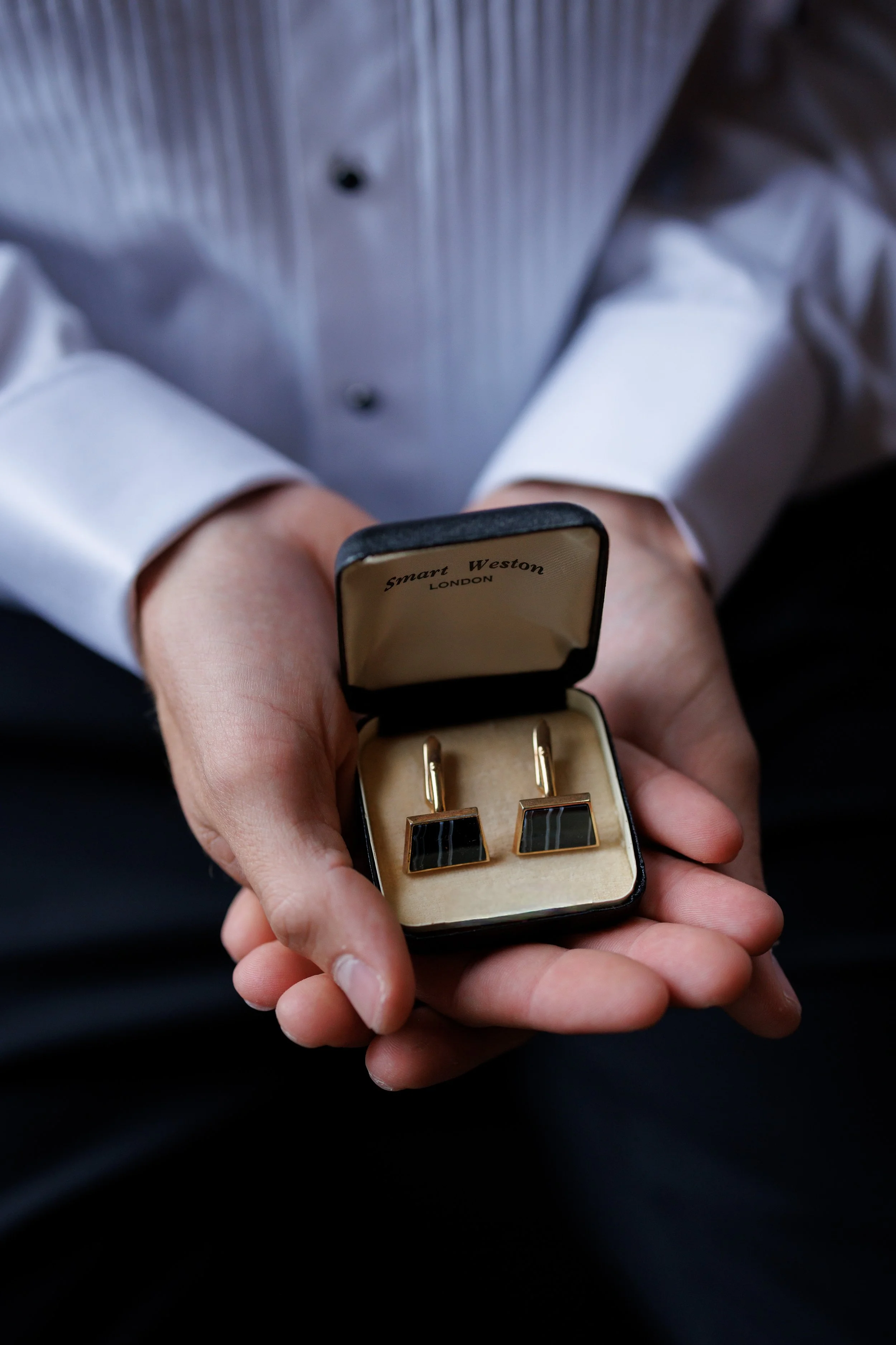 A person holding an open jewelry box containing a pair of black and gold cufflinks.