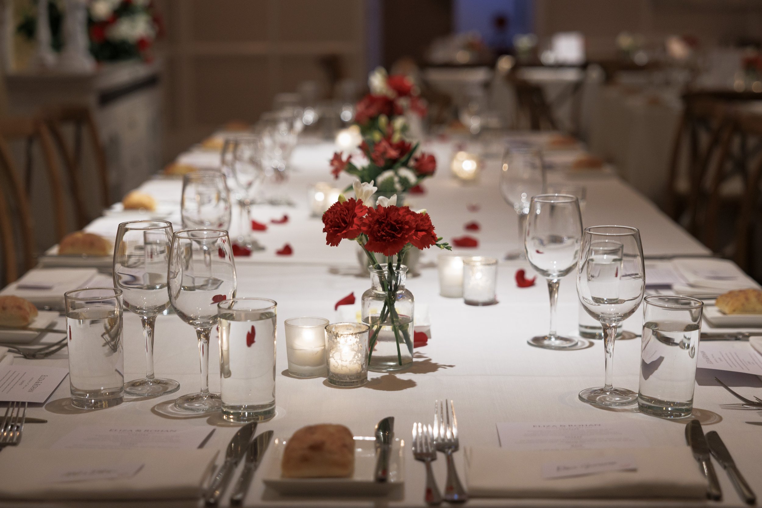 Elegant dining table decorated with red and white flowers, candles, glasses, and silverware, set for a formal event.