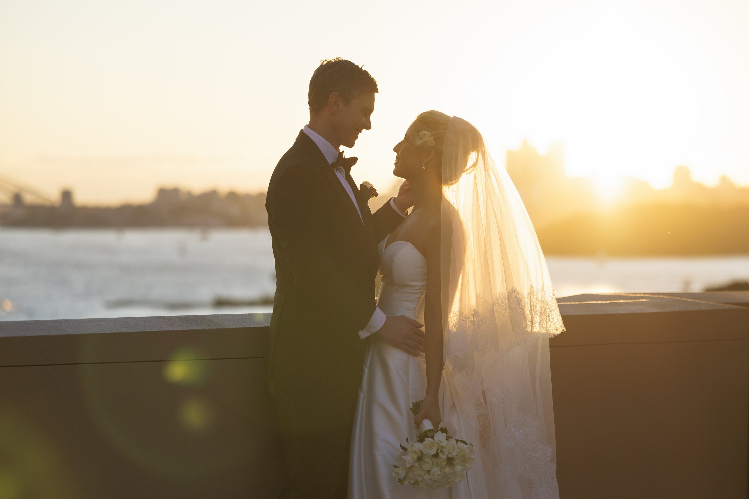 A newlywed couple in formal wedding attire standing close together at sunset near a body of water.