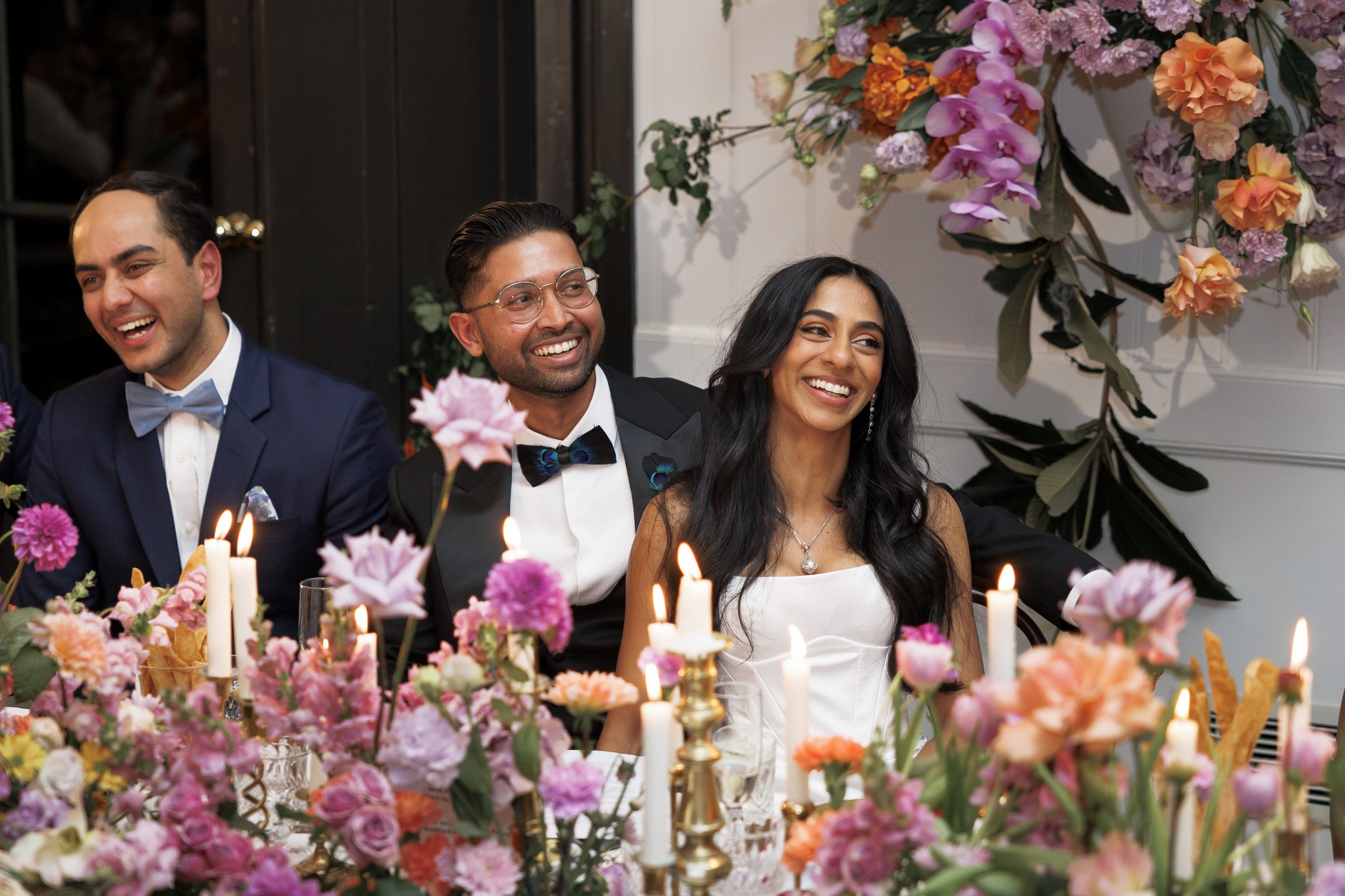People dressed in formal attire, smiling and enjoying a celebration with floral decorations and candles.