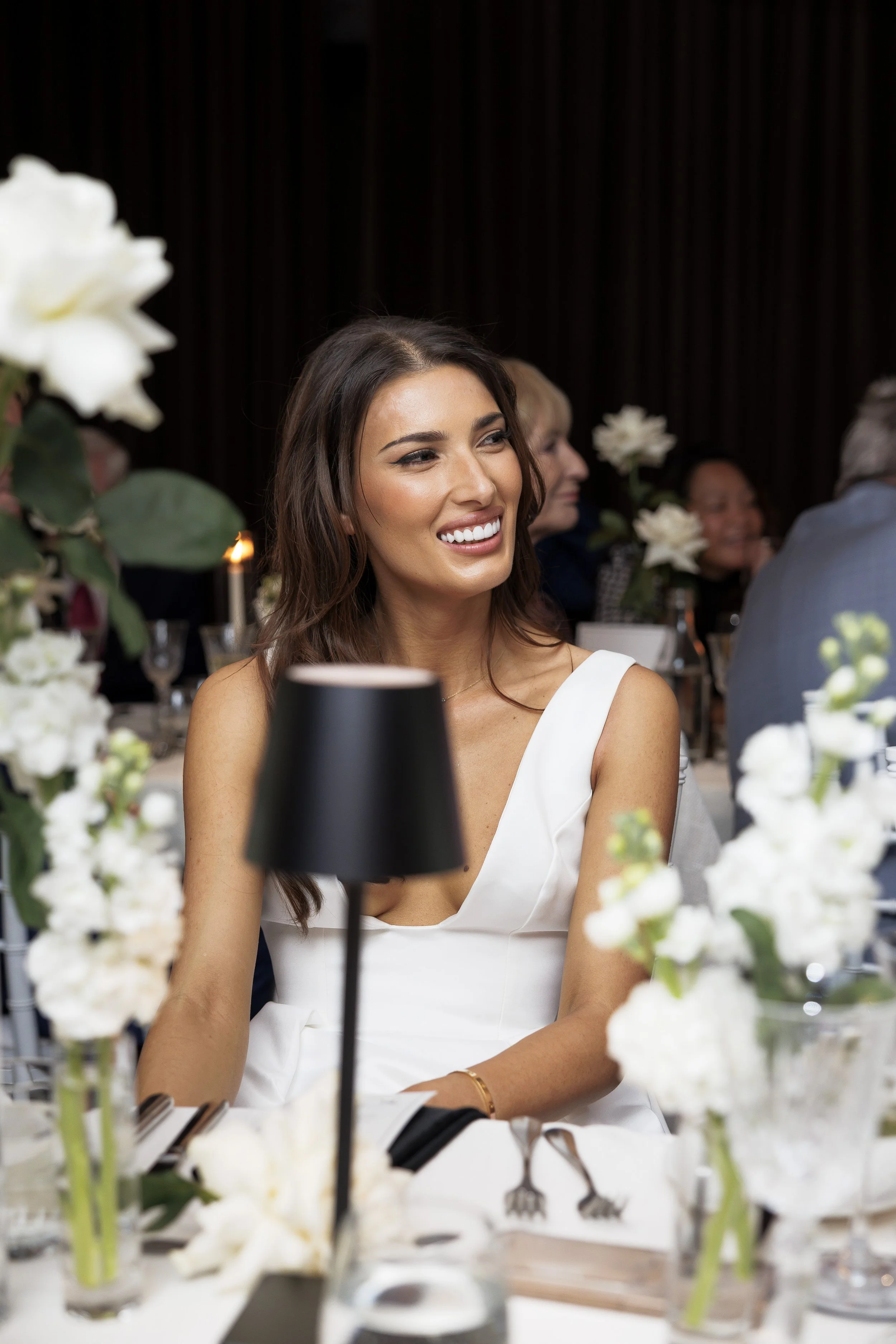 A woman with long brown hair, wearing a white dress, sitting at a table during a formal event with other guests, surrounded by white flowers and black table lamps, smiling and looking to her right.
